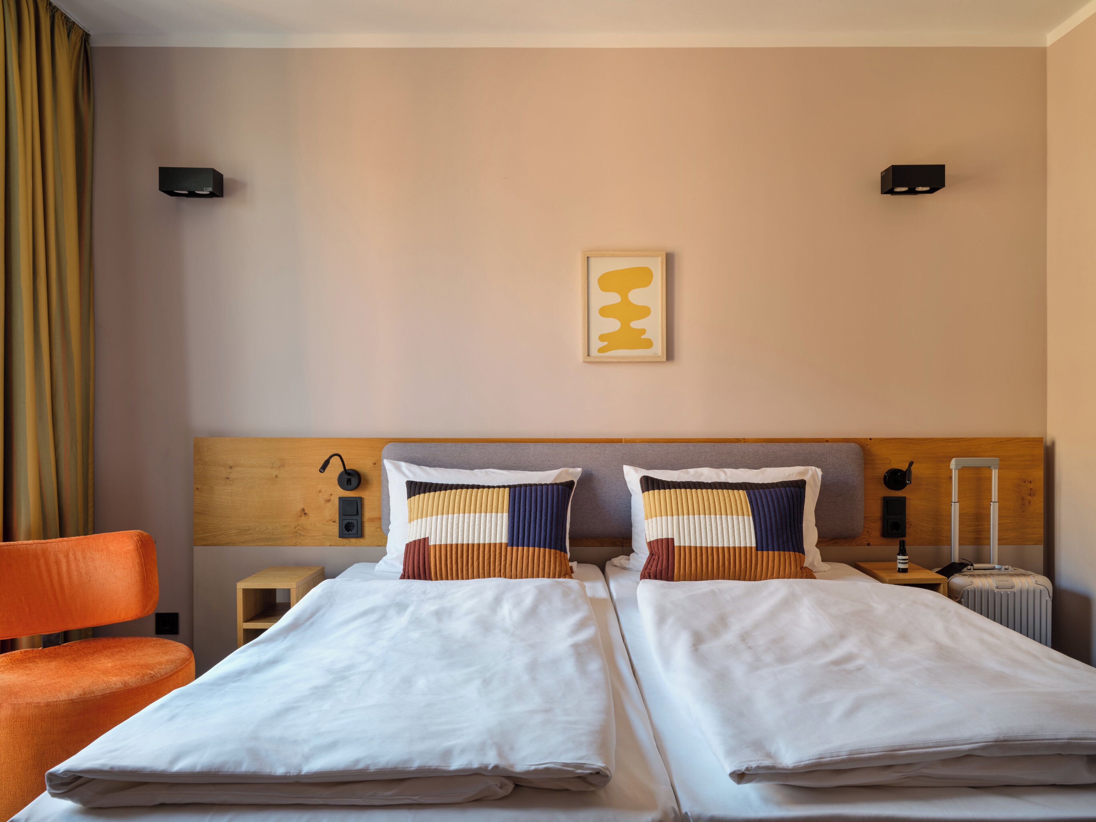 Ein Hotelzimmer mit zwei Einzelbetten, einem Holzkopfteil, einem kleinen Nachttisch mit Lampe, einer Steckdose und einem Koffer auf dem Boden.