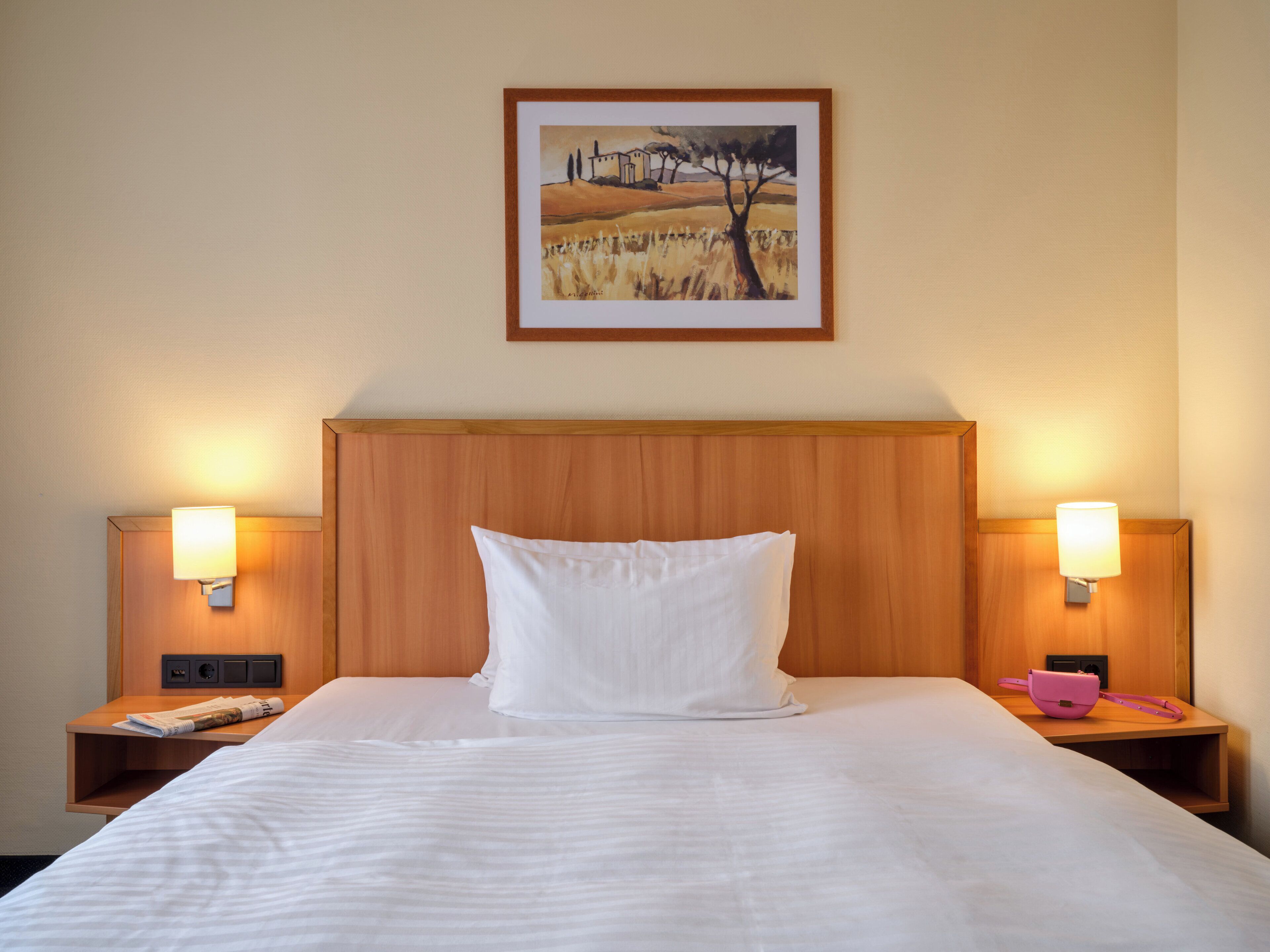 Ein Hotelzimmer mit einem großen Bett, zwei Nachttischen mit Lampen, einem gerahmten Gemälde an der Wand und einem hölzernen Kopfteil.