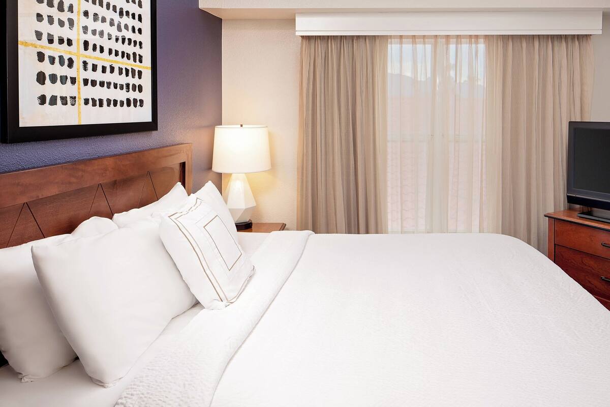 A hotel room with a large bed, a TV on a wooden stand, a lamp, and a window with curtains.