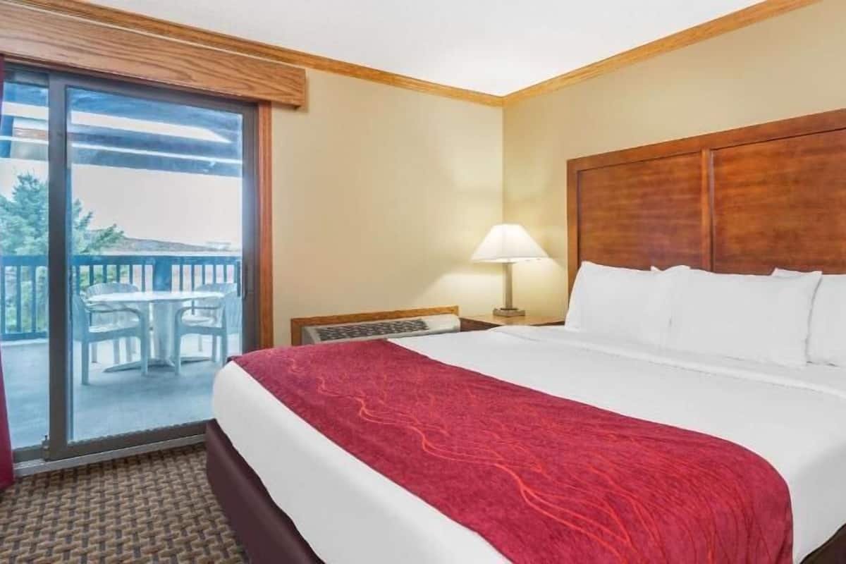 A hotel room with a large bed, a balcony with a table and chairs, a nightstand with a lamp, and a wooden headboard.