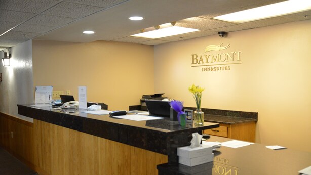 A hotel reception desk with a wooden counter, a computer, and a vase with flowers. - Baymont by Wyndham Kasson Rochester Area (Kasson)