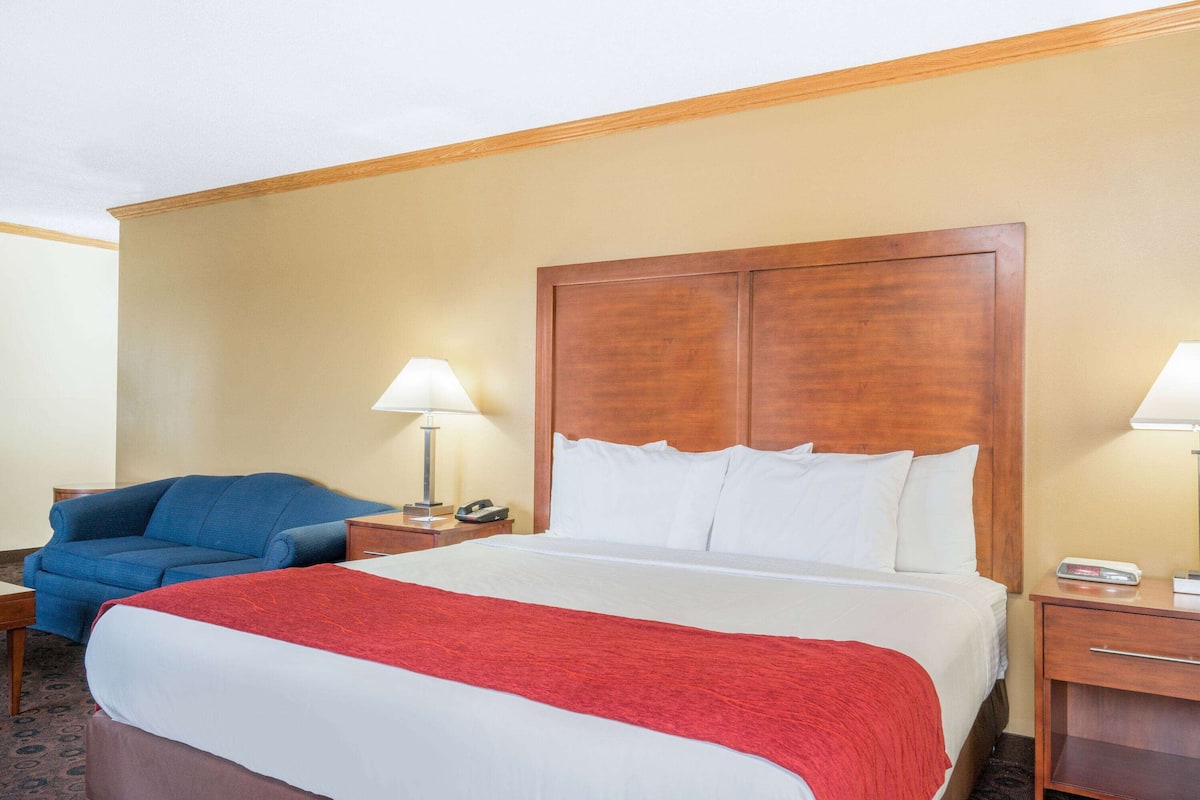 A hotel room with a large bed, a blue armchair, two bedside tables with lamps, and a wooden headboard.