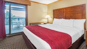 A hotel room with a large bed, a view through a sliding glass door to an outdoor area, a nightstand with a lamp, and a wooden headboard. - Baymont by Wyndham Kasson Rochester Area (Kasson)