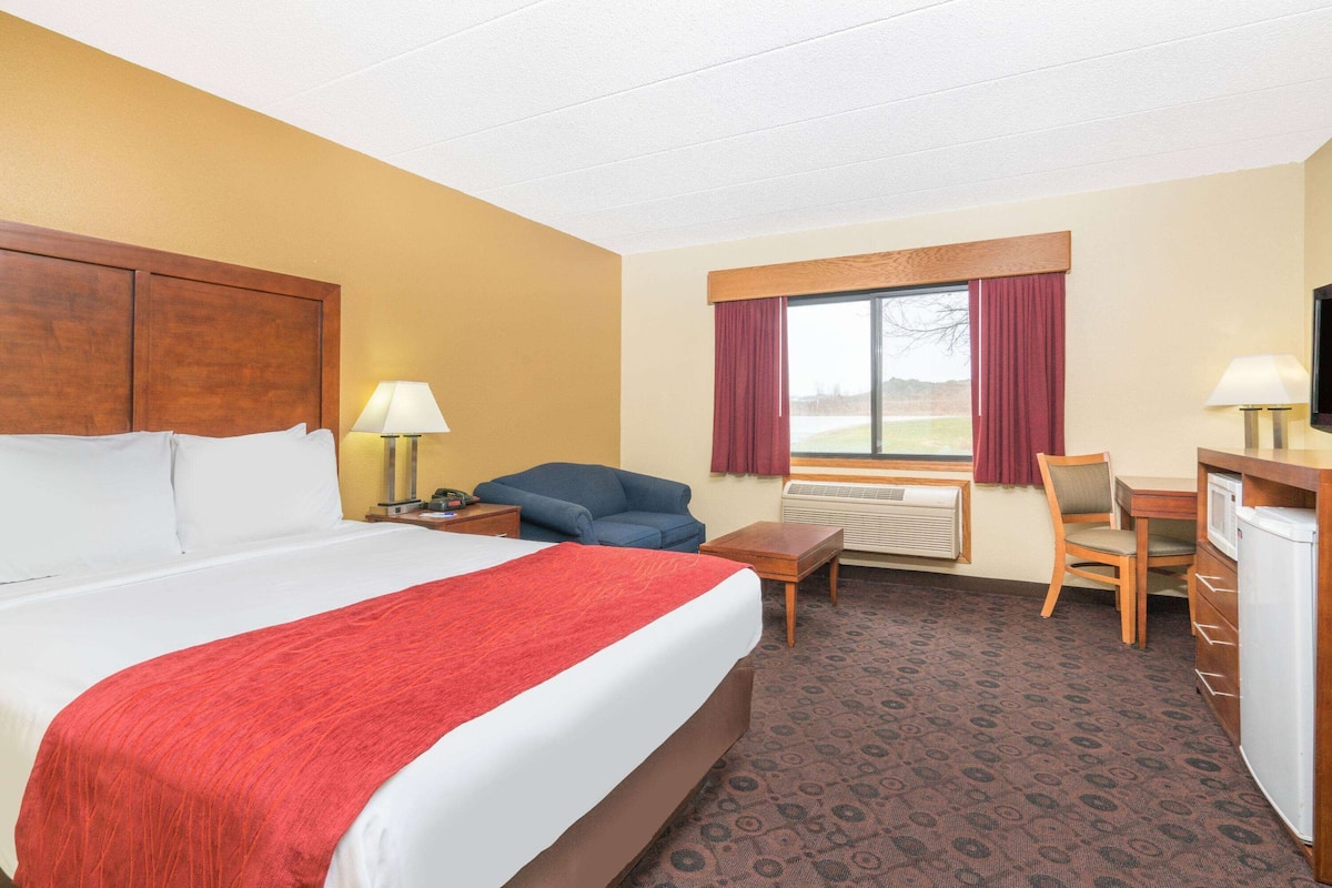 A hotel room with a large bed, a desk, a chair, a small table, a lamp, and a television.