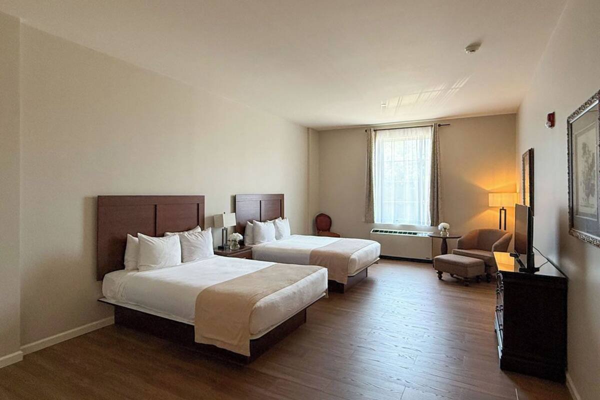 A hotel room with two beds, a desk, a chair, a lamp, and a window with curtains.