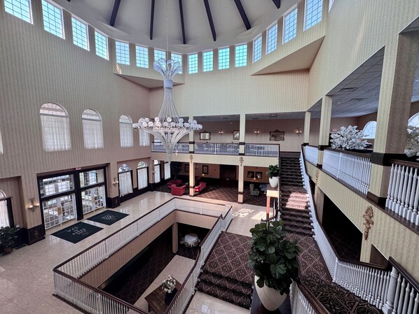 A spacious hotel lobby with multiple levels, a central chandelier, and a staircase leading to the upper floor.