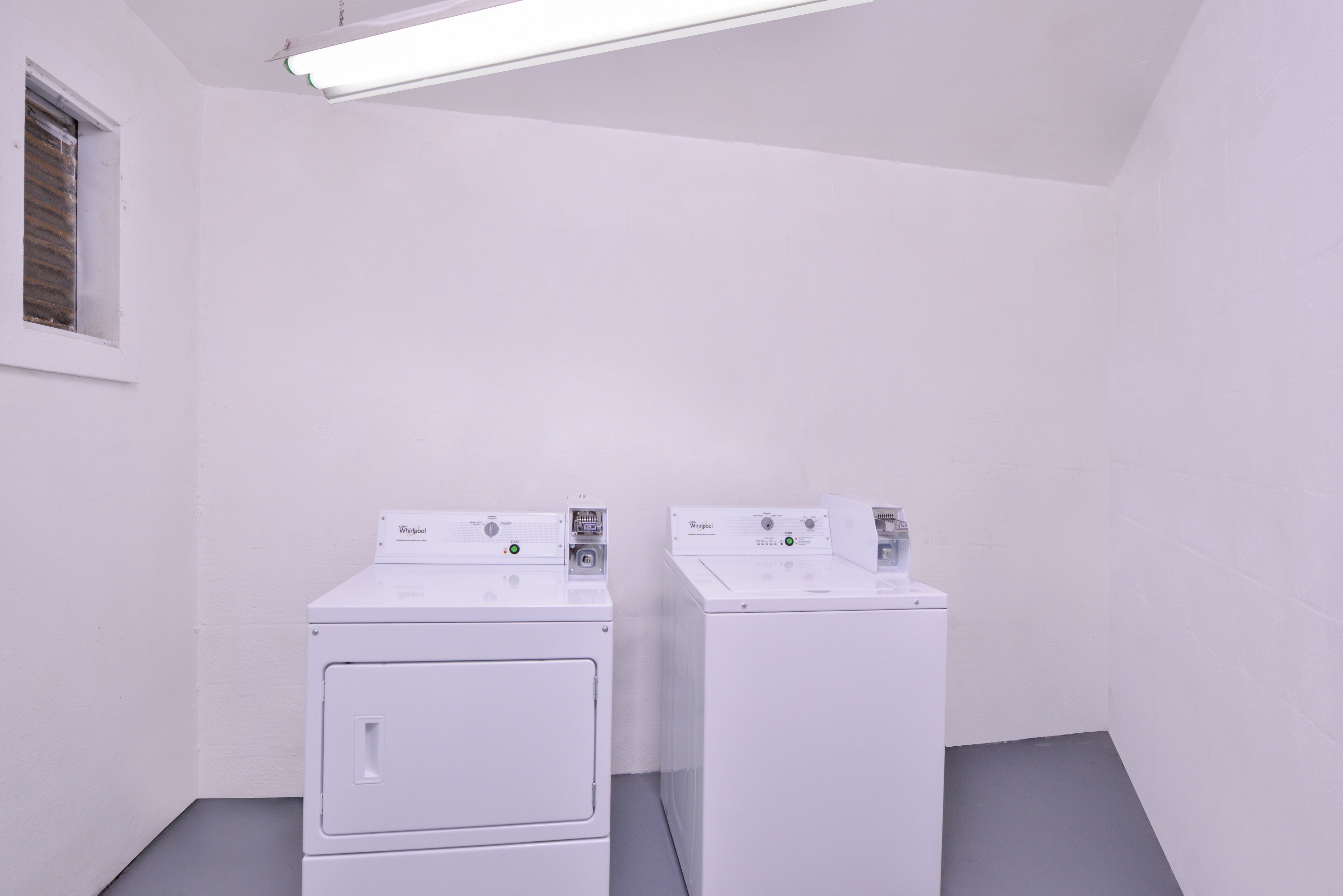 A laundry room with two white Whirlpool washers and dryers, a small window, and a fluorescent light fixture.