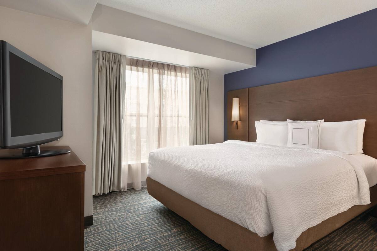 A hotel room with a large bed, a flat-screen TV on a wooden stand, a window with sheer curtains, and a wall-mounted lamp.