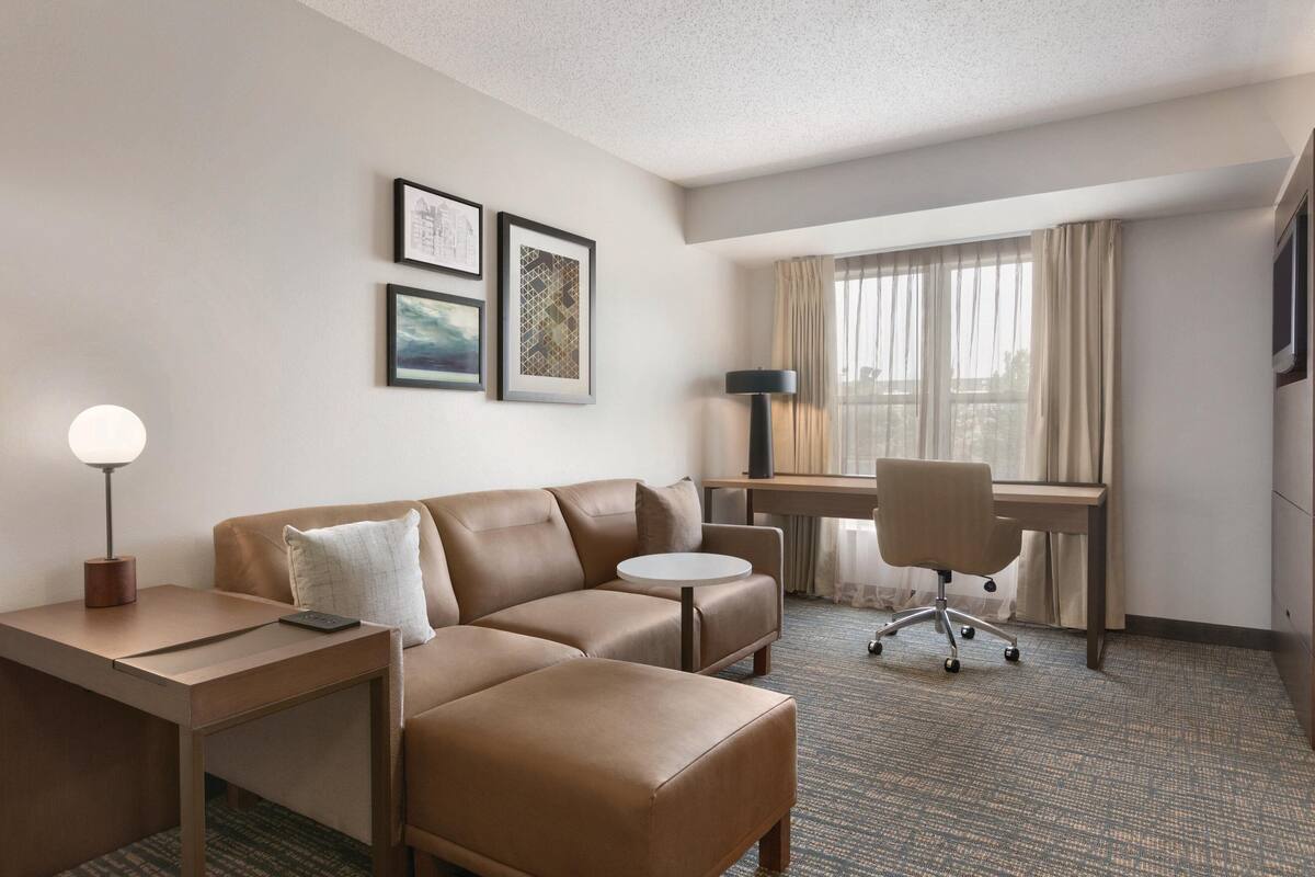 A hotel room with a brown leather sofa, a small round table, a desk with a chair, and a lamp. There are framed pictures on the wall and a window with curtains.
