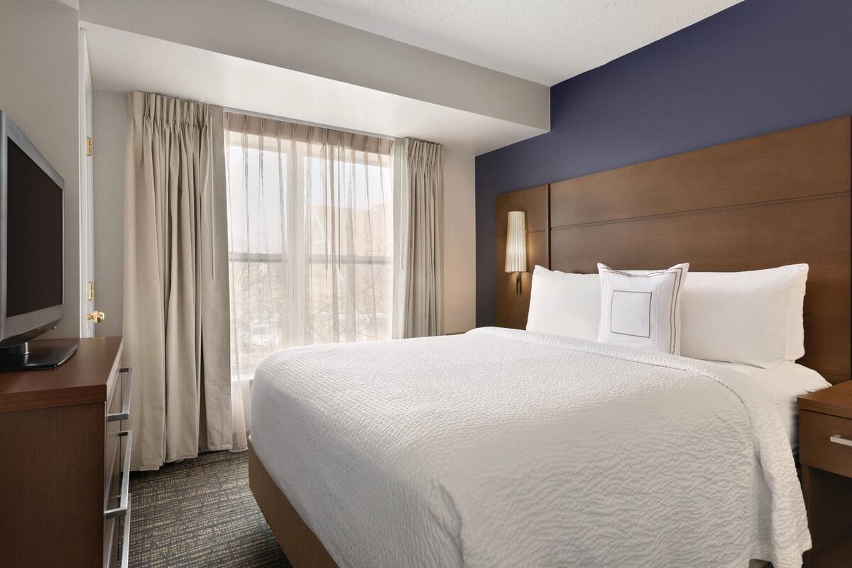 A hotel room with a large bed, a flat-screen TV, a wooden headboard, and a window with curtains.