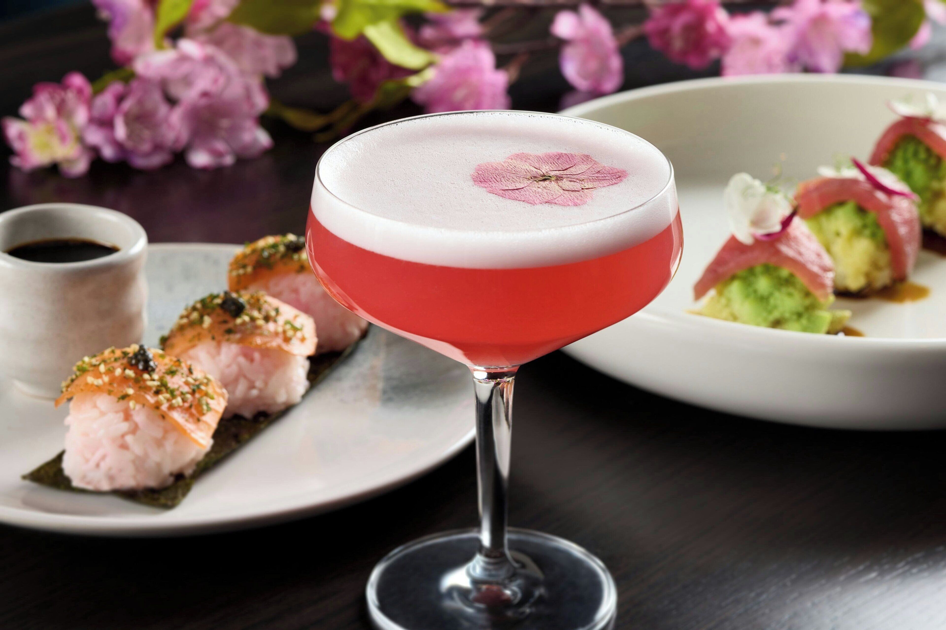 A cocktail in a martini glass with a pink and white color scheme, accompanied by sushi on a plate.