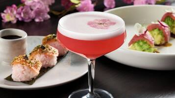 A cocktail in a martini glass with a pink and white color scheme, accompanied by sushi on a plate.