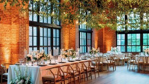 A long dining table set with candles, flowers, and chairs in a room with brick walls and large windows.