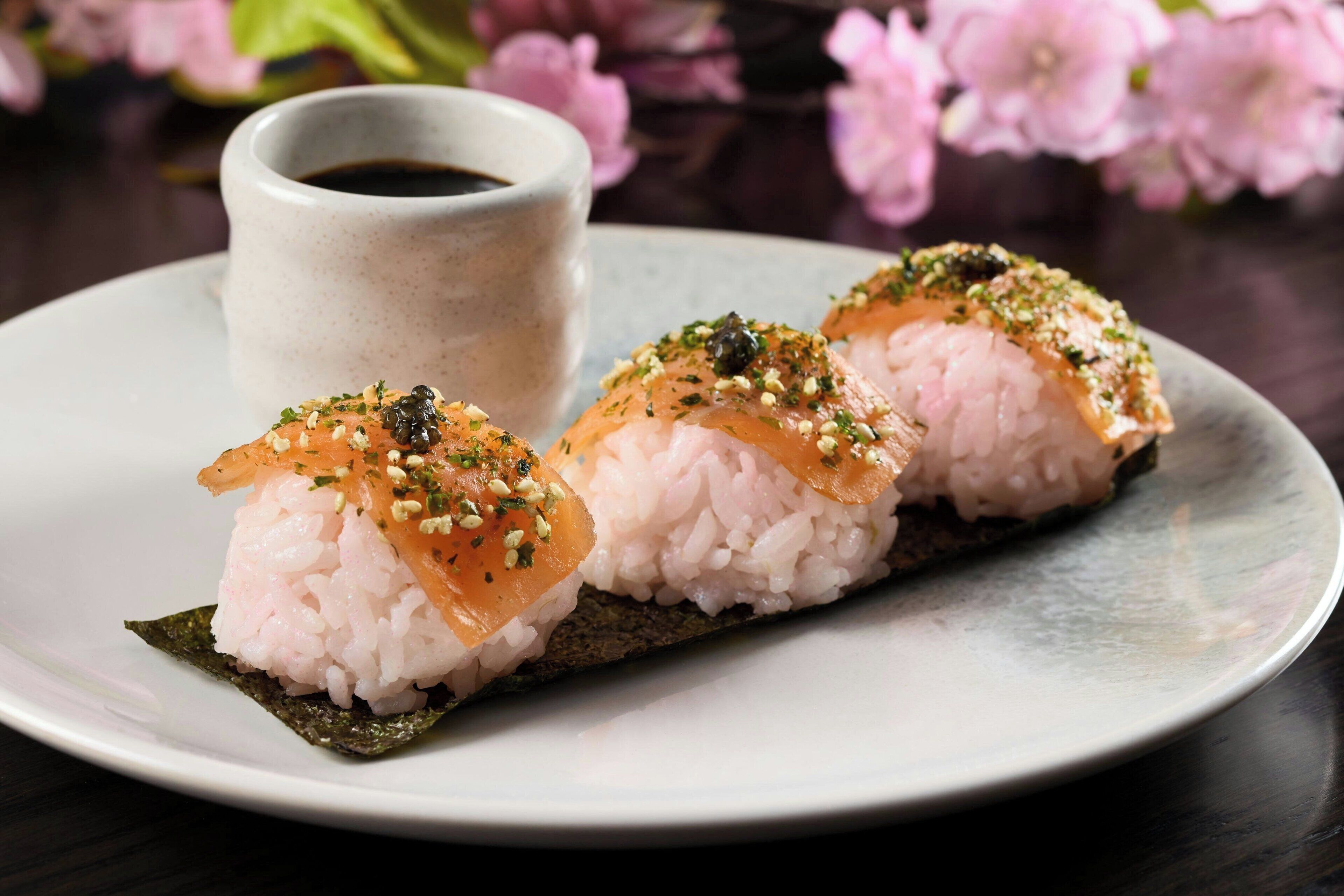Ein Teller mit drei Sushi-Stücken, einer kleinen Sojasaucenschale und rosa Blumen im Hintergrund.