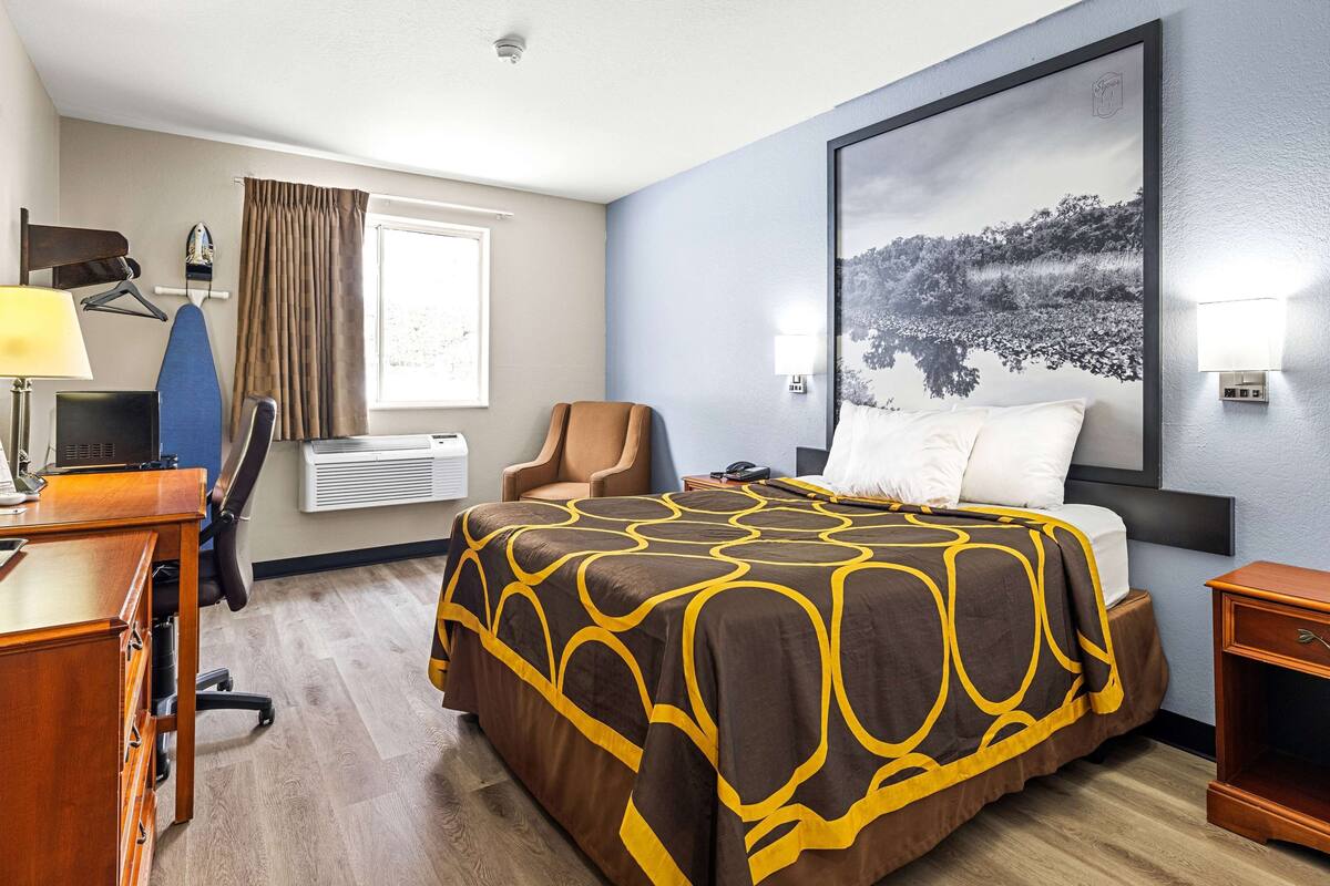 A hotel room with a bed, a desk, a chair, an ironing board, and a window with curtains.