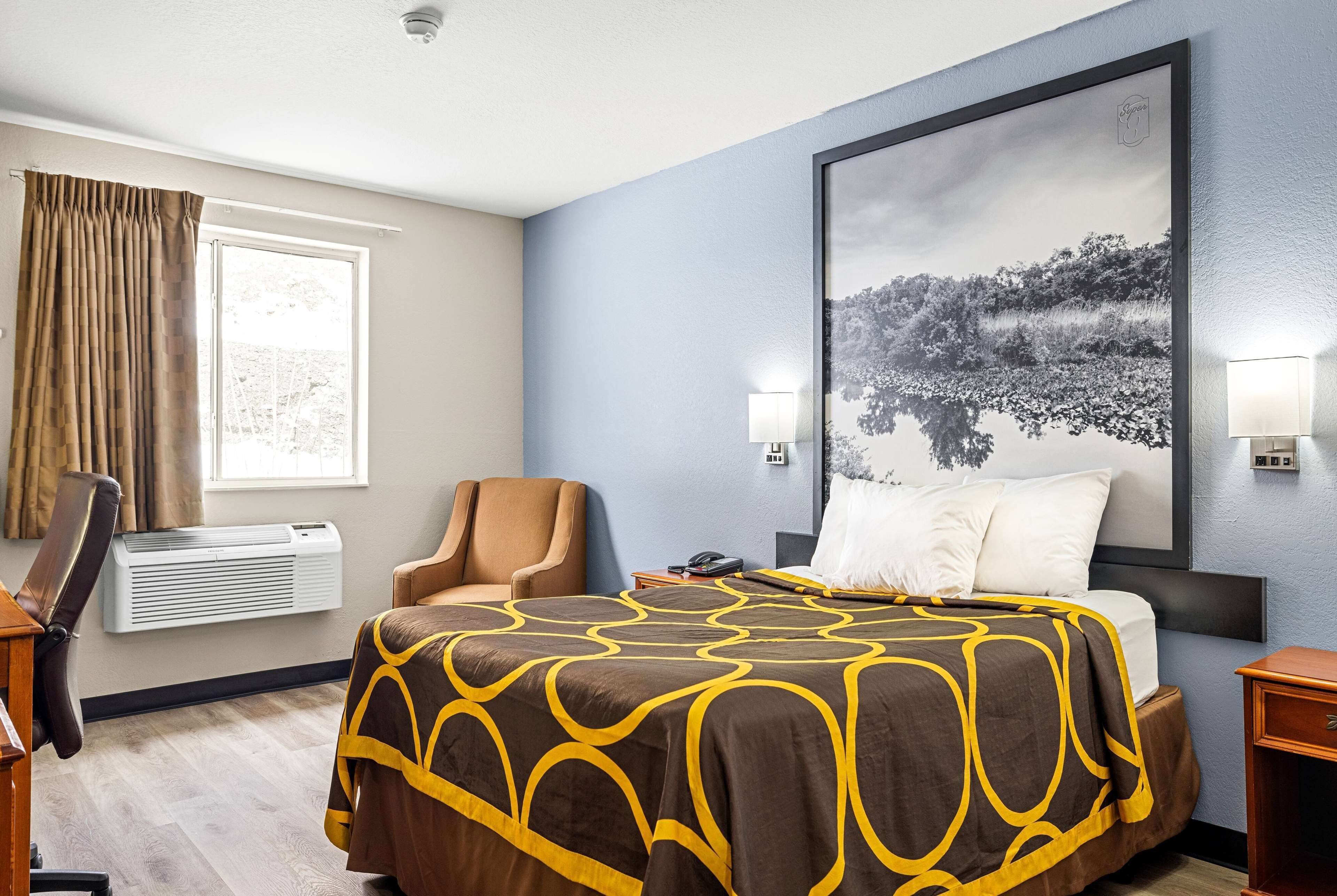 A hotel room with a bed, a chair, a desk, an air conditioner, a window with curtains, and a wall-mounted picture.
