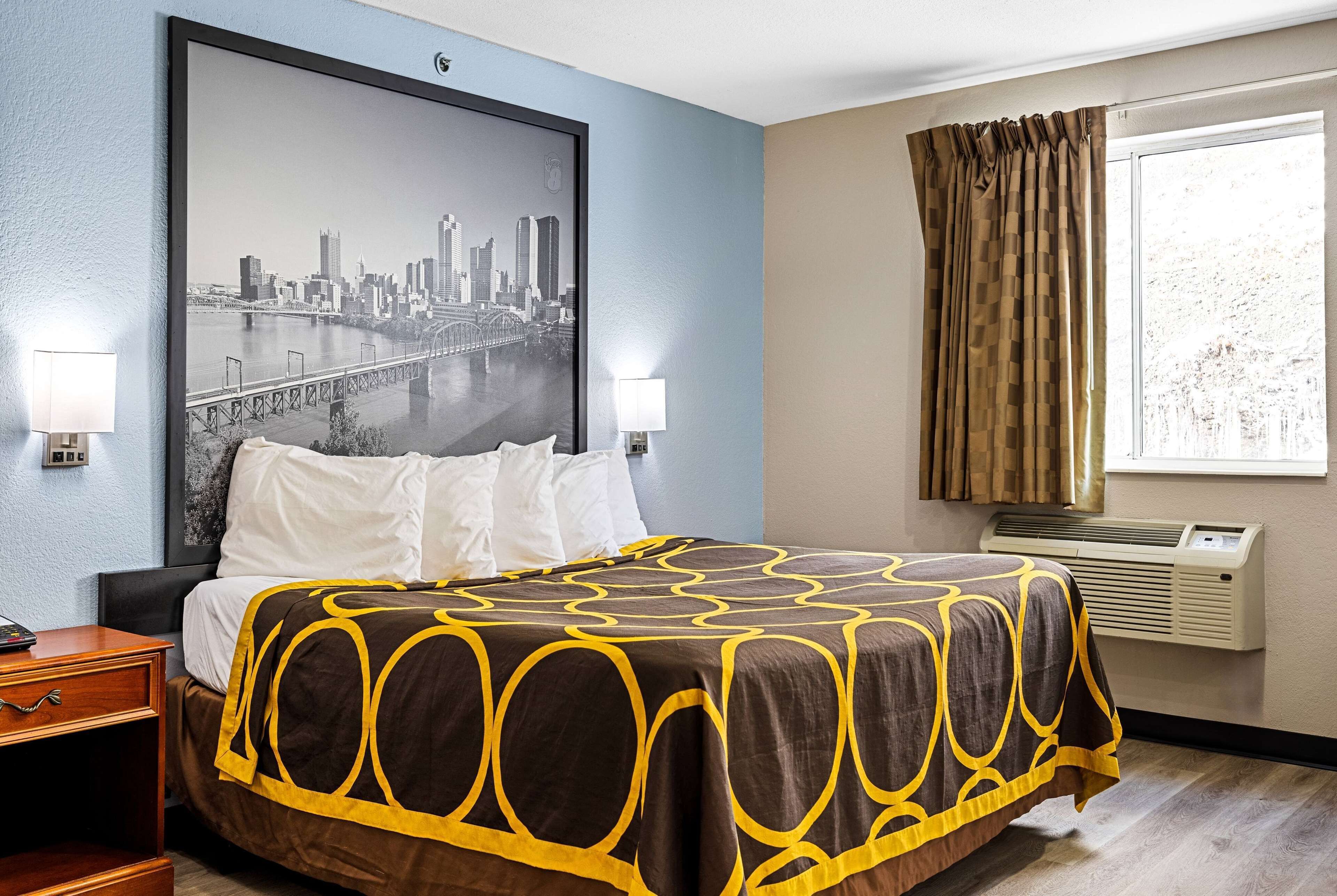 A hotel room with a large bed, two bedside lamps, a cityscape poster on the wall, and a window with curtains.
