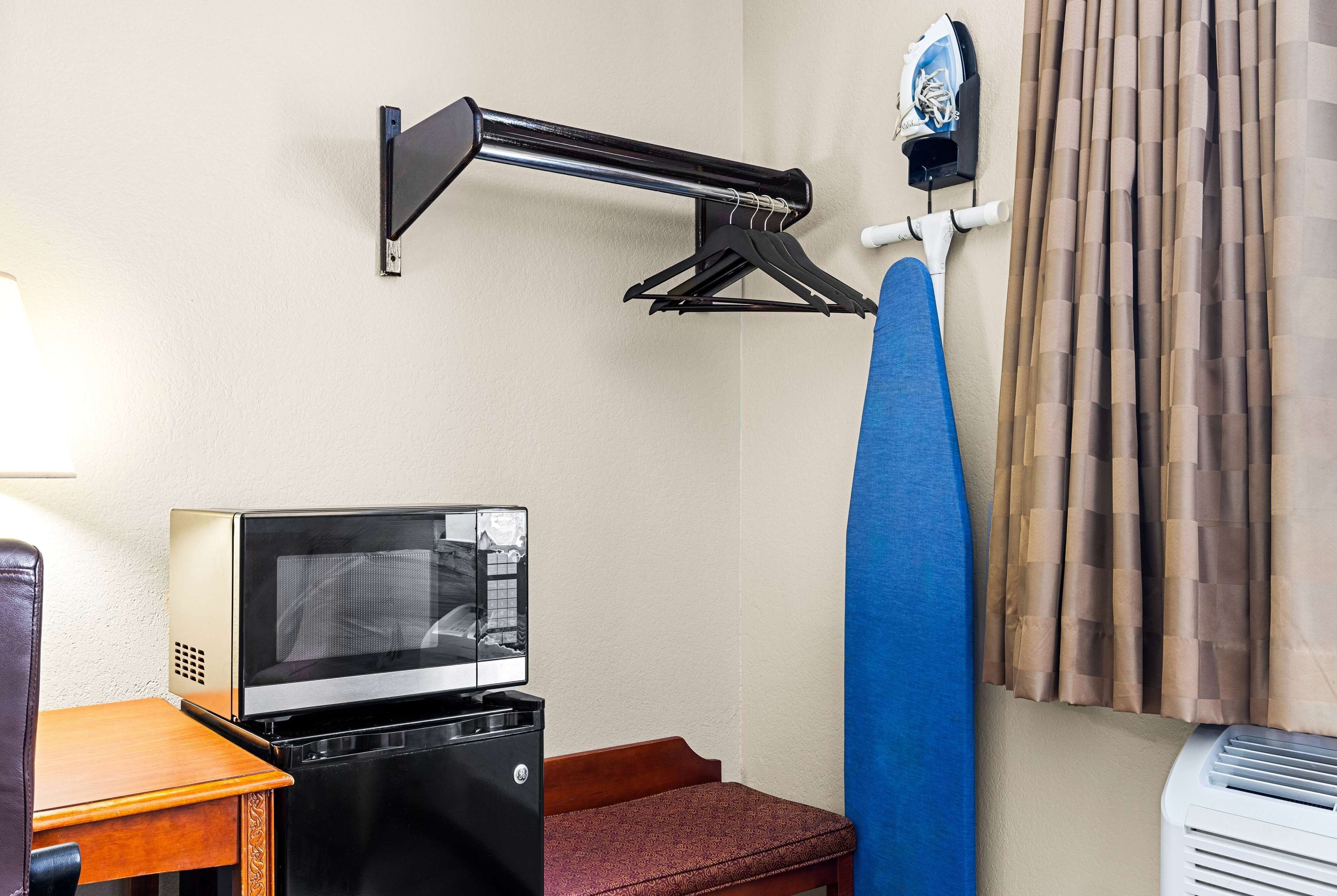 A compact hotel room with a television, a small table, a bench, a wall-mounted ironing board, and a window with curtains.