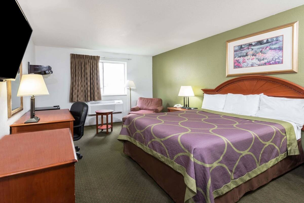 A hotel room with a bed, desk, chair, and a painting on the wall.