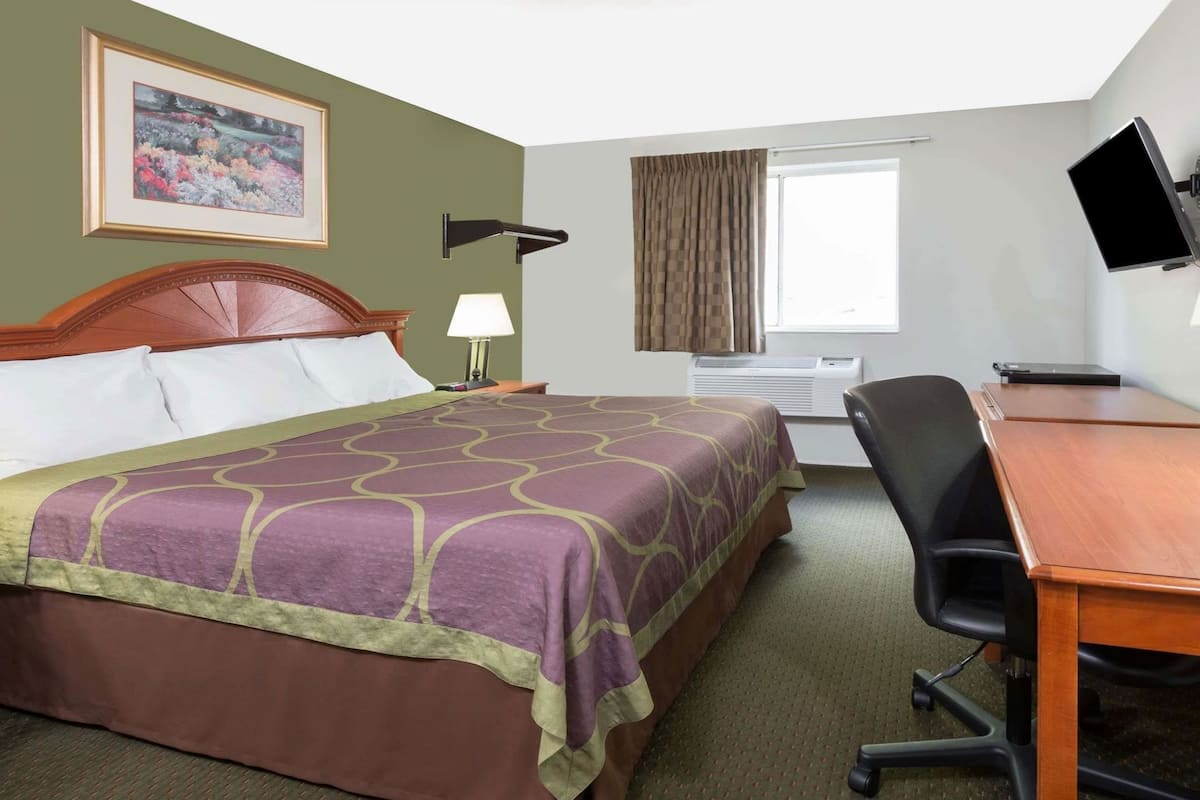 A hotel room with a bed, desk, chair, TV, air conditioning unit, and a window with curtains.