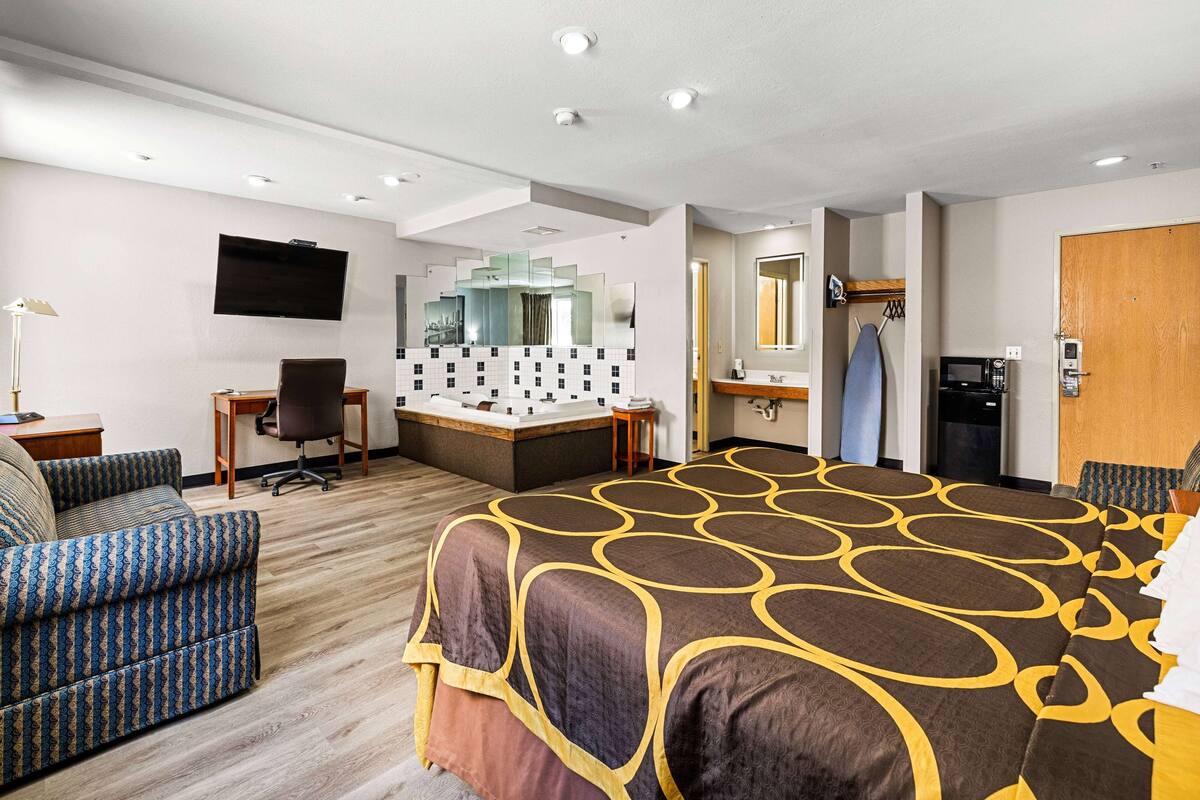 A hotel room with a large bed, a sofa, a desk with a computer, a TV, a bathroom with a mirror, and a microwave.