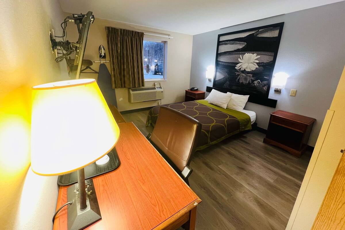 A hotel room with a wooden desk, a bed with a patterned bedspread, a wall-mounted air conditioner, a window with curtains, and a large floral painting on the wall.