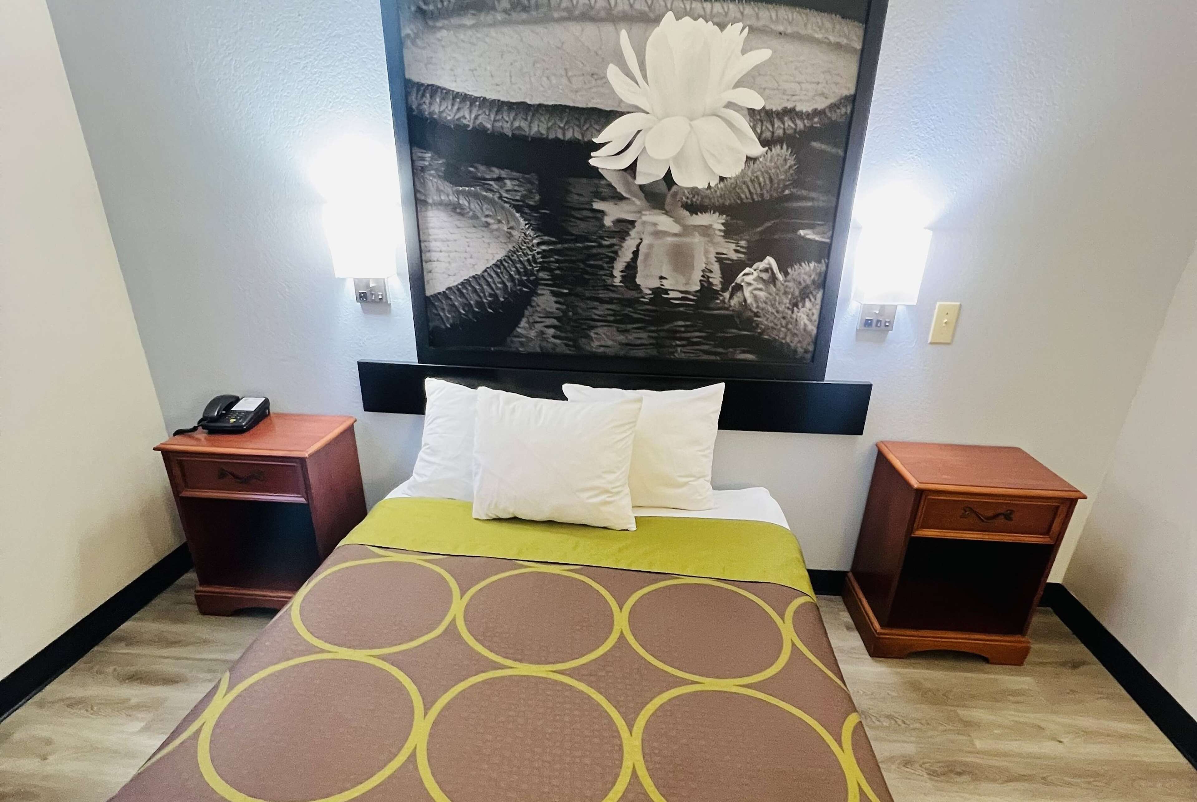 Une chambre d’hôtel avec un lit, une table de chevet avec un téléphone, une lampe fixée au mur et une grande œuvre d’art florale au-dessus du lit.