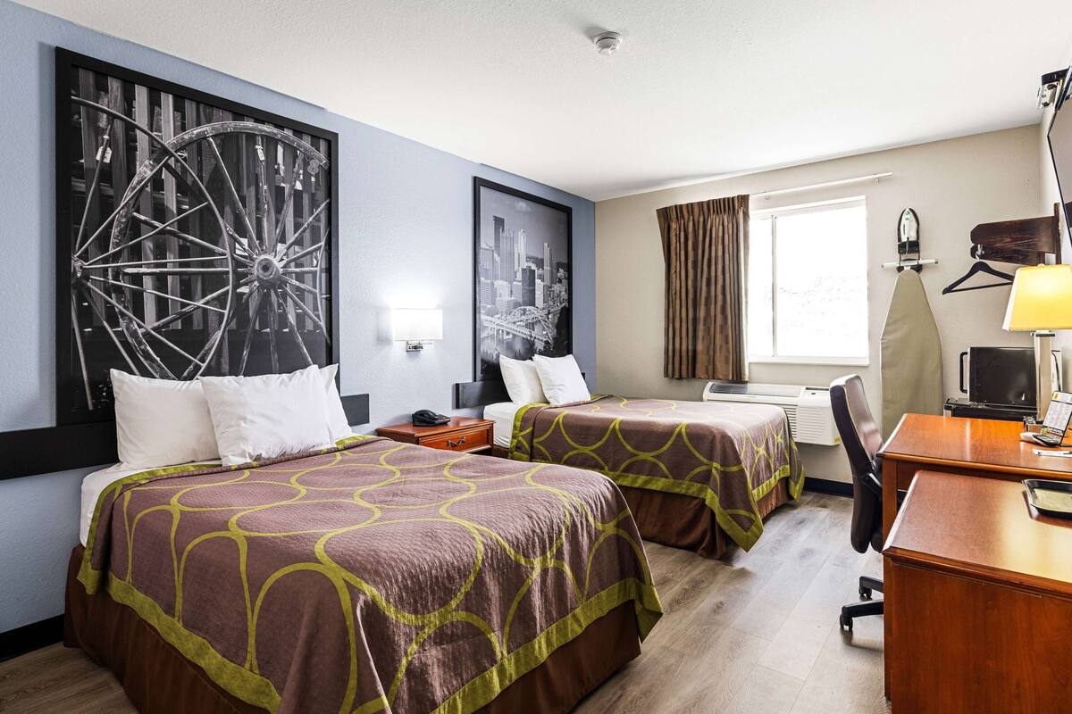 A hotel room with two beds, a desk, a chair, a large window with curtains, and artwork on the walls.