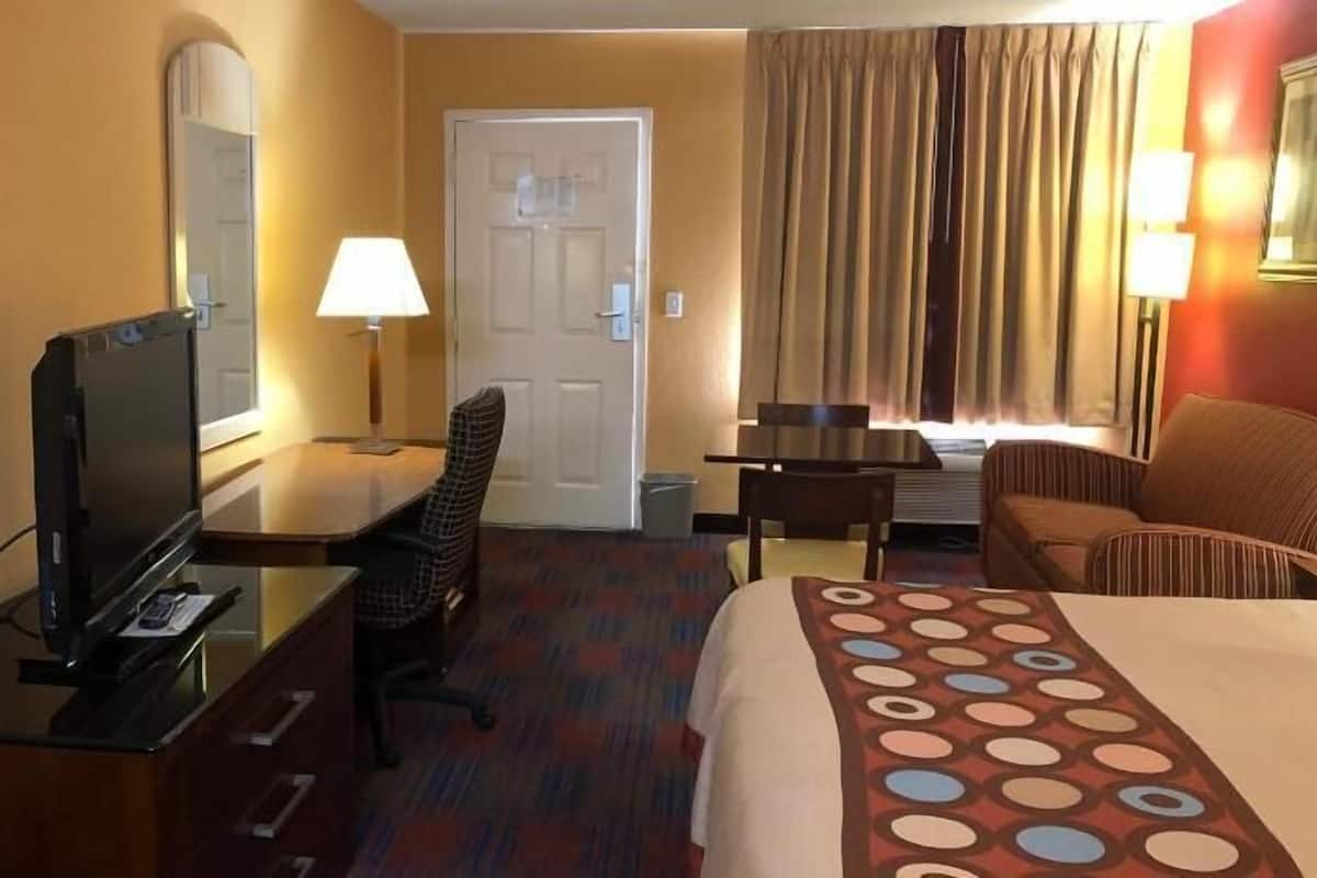 A hotel room with a bed, a desk with a television, a chair, a lamp, a door, and a window with curtains.