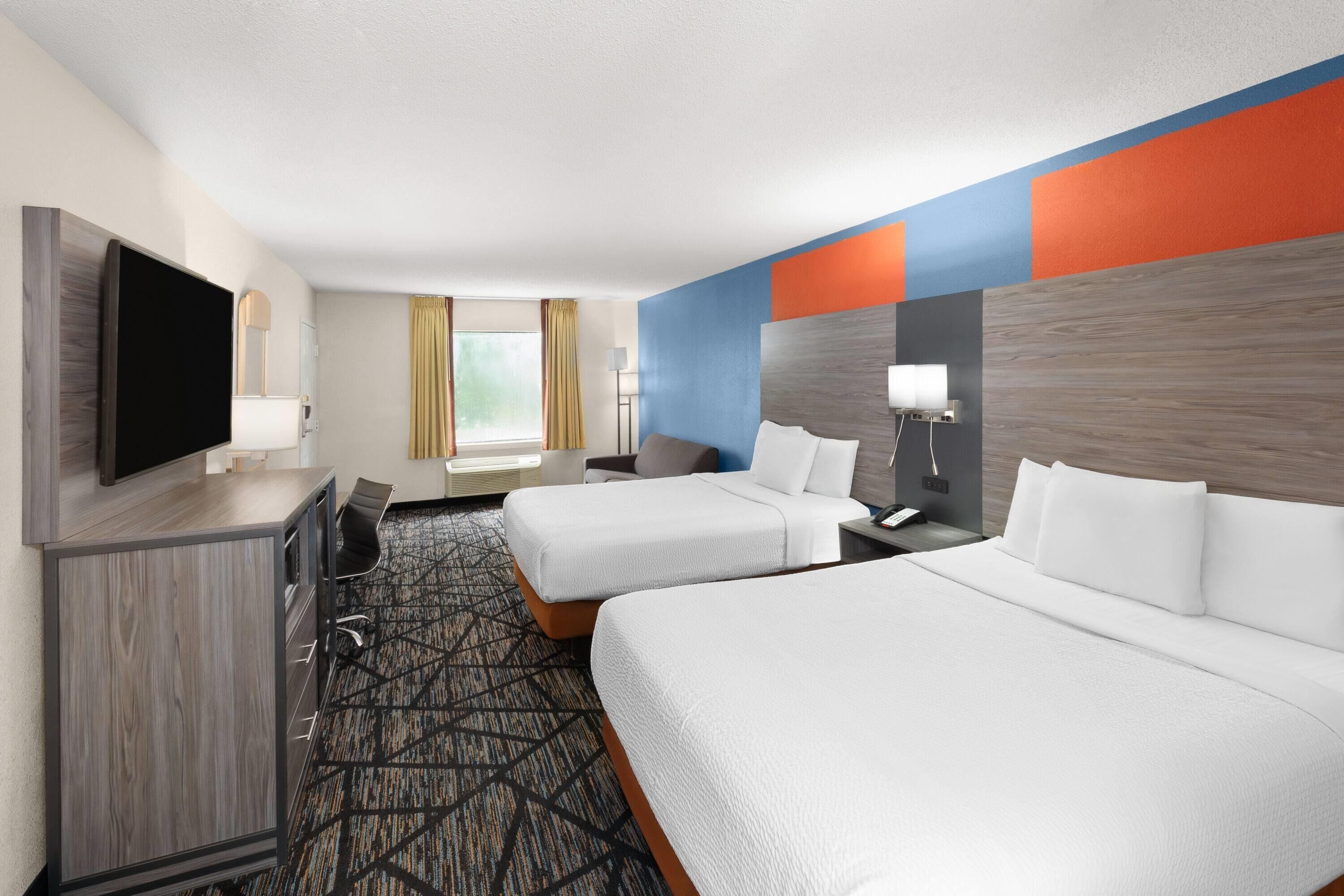 Quarto de hotel com duas camas, uma escrivaninha, televisão e janela com cortinas.