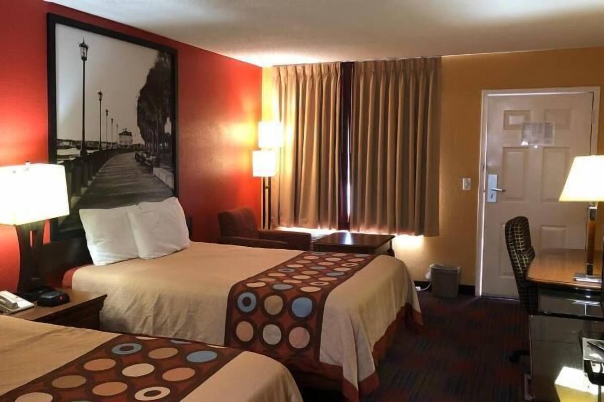 A hotel room with two beds, a desk, a chair, a lamp, and a painting on the wall.