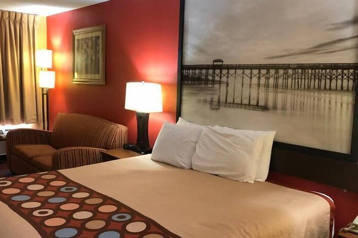 A hotel room with a bed, a chair, a desk, a lamp, a painting, and a telephone.