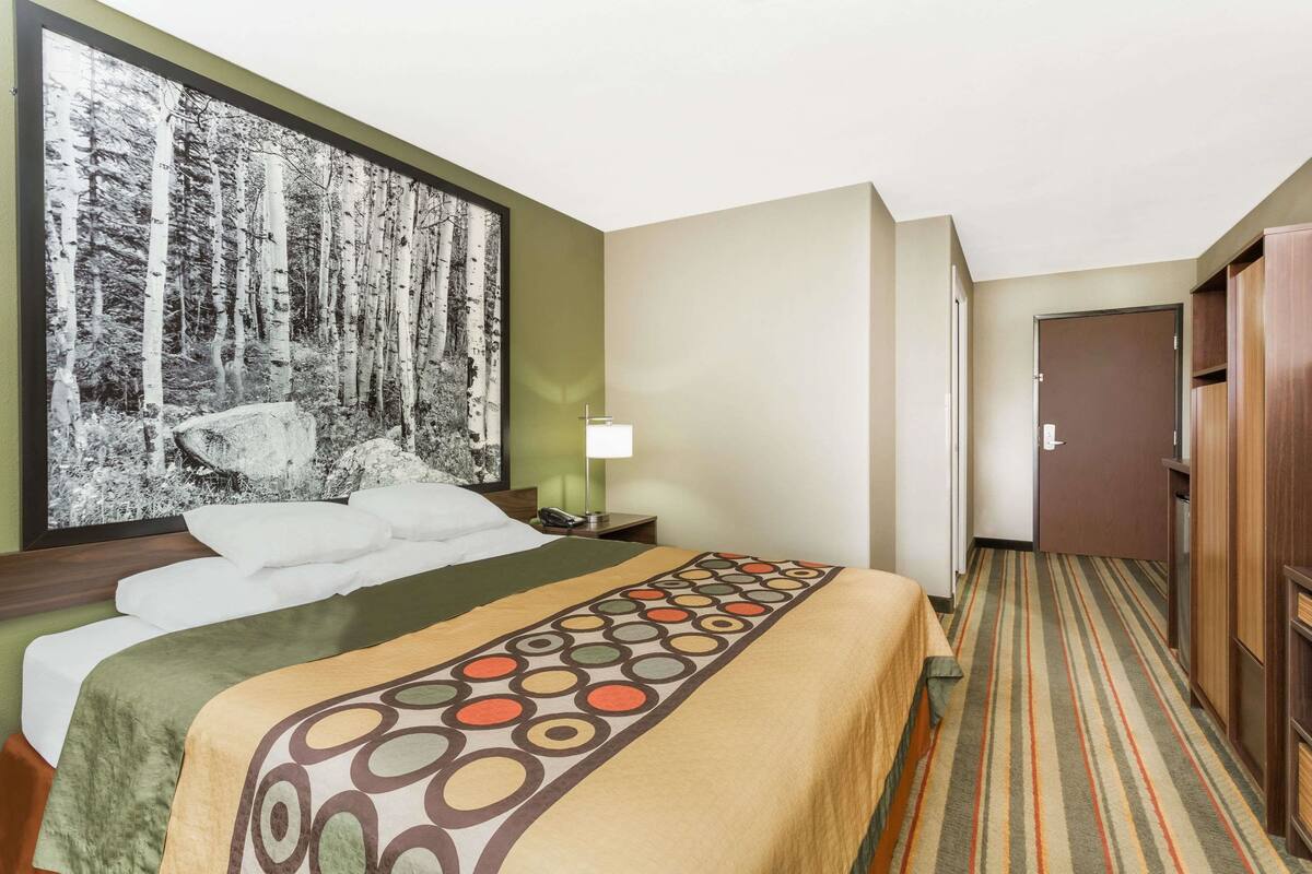 Ein Hotelzimmer mit einem großen Bett, einem Nachttisch mit Lampe, einem Kleiderschrank und einem gerahmten Foto von einem Wald an der Wand.