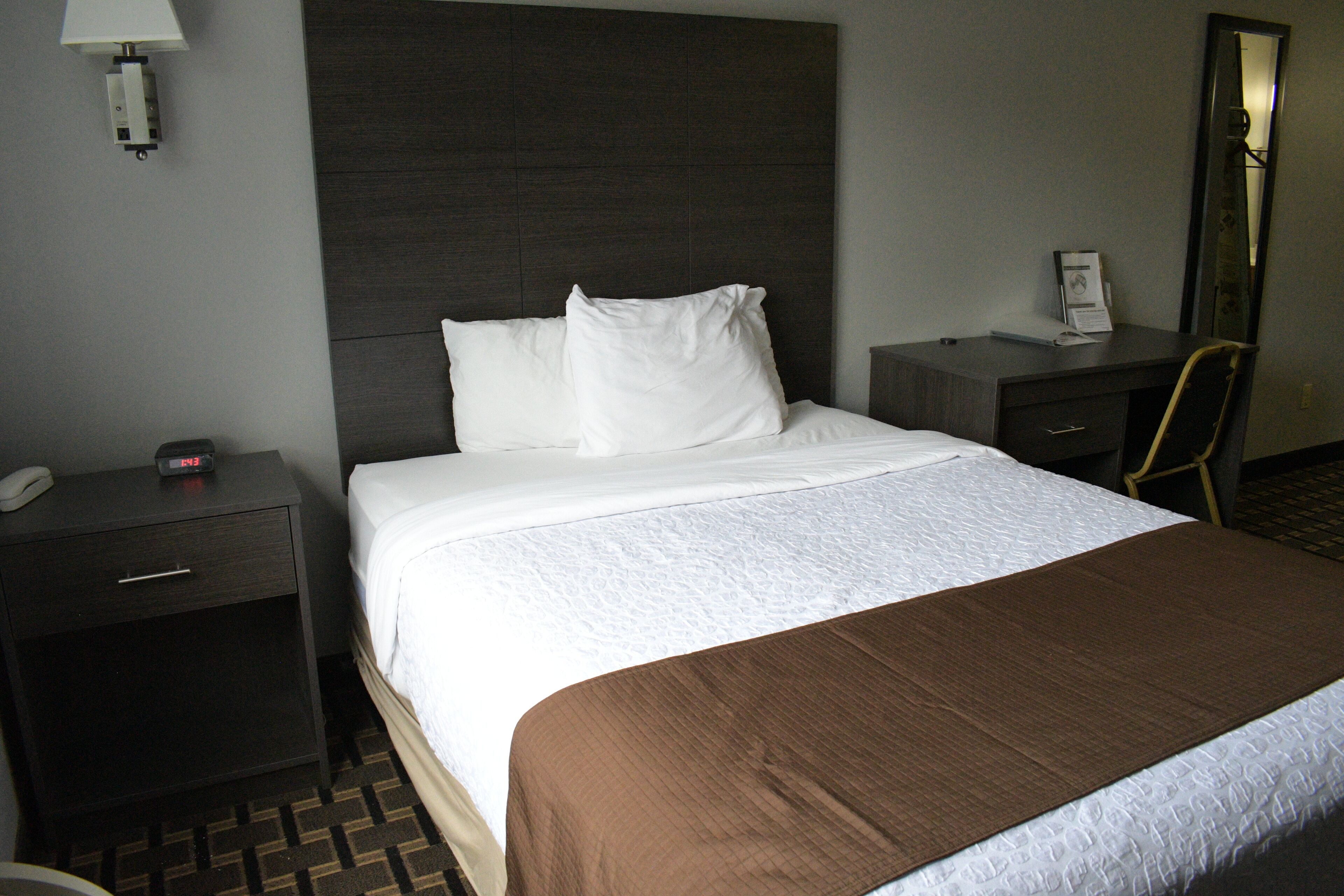 A hotel room with a large bed, a nightstand with a phone and a clock, a desk with a chair, and a mirror on the wall.