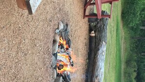 A campfire with logs and rocks arranged in a circle, surrounded by a gravel area with a wooden structure and a red bench in the background. - Adams Inn (Adams)
