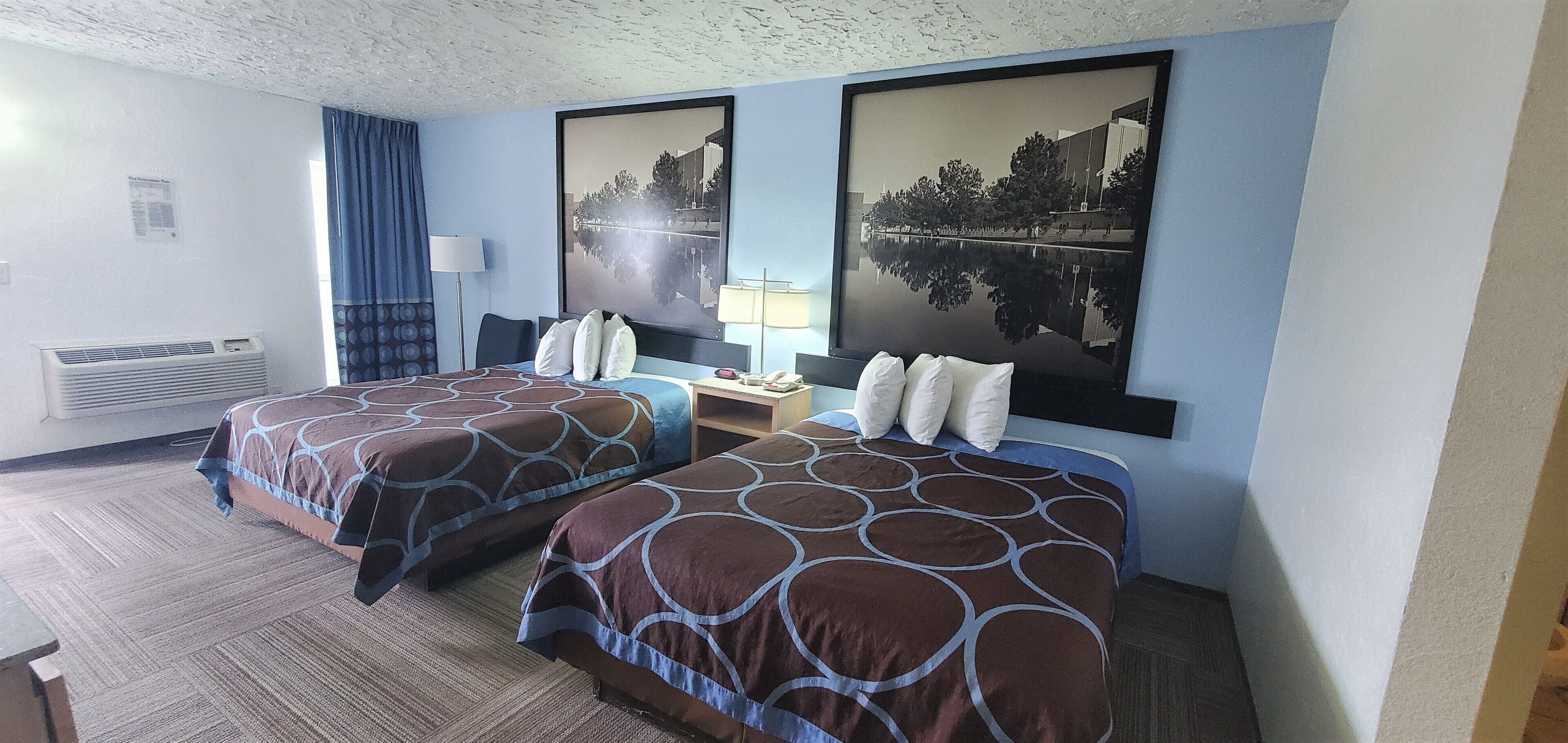 A hotel room with two beds, a nightstand, a lamp, and a large window with blue curtains.