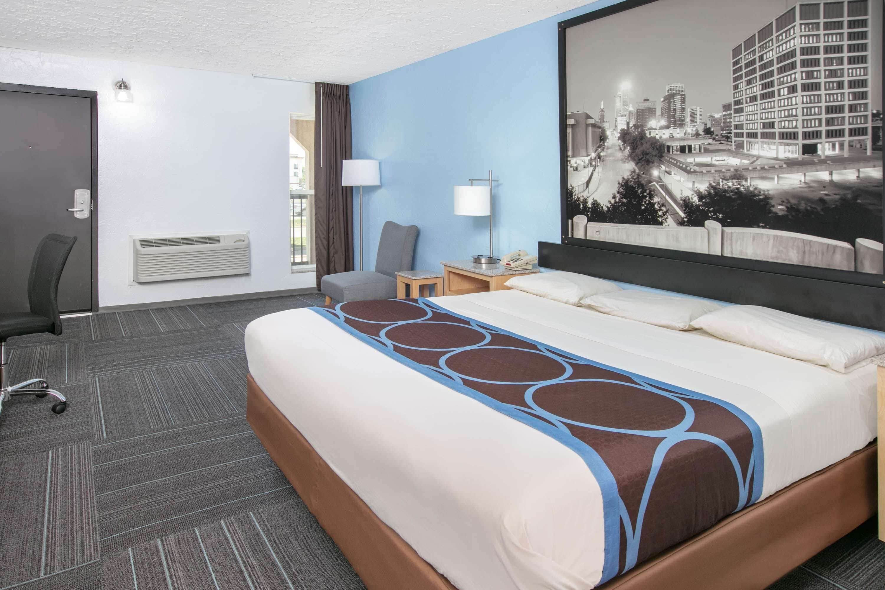 A hotel room with a large bed, a desk, a chair, a lamp, and a cityscape painting on the wall.