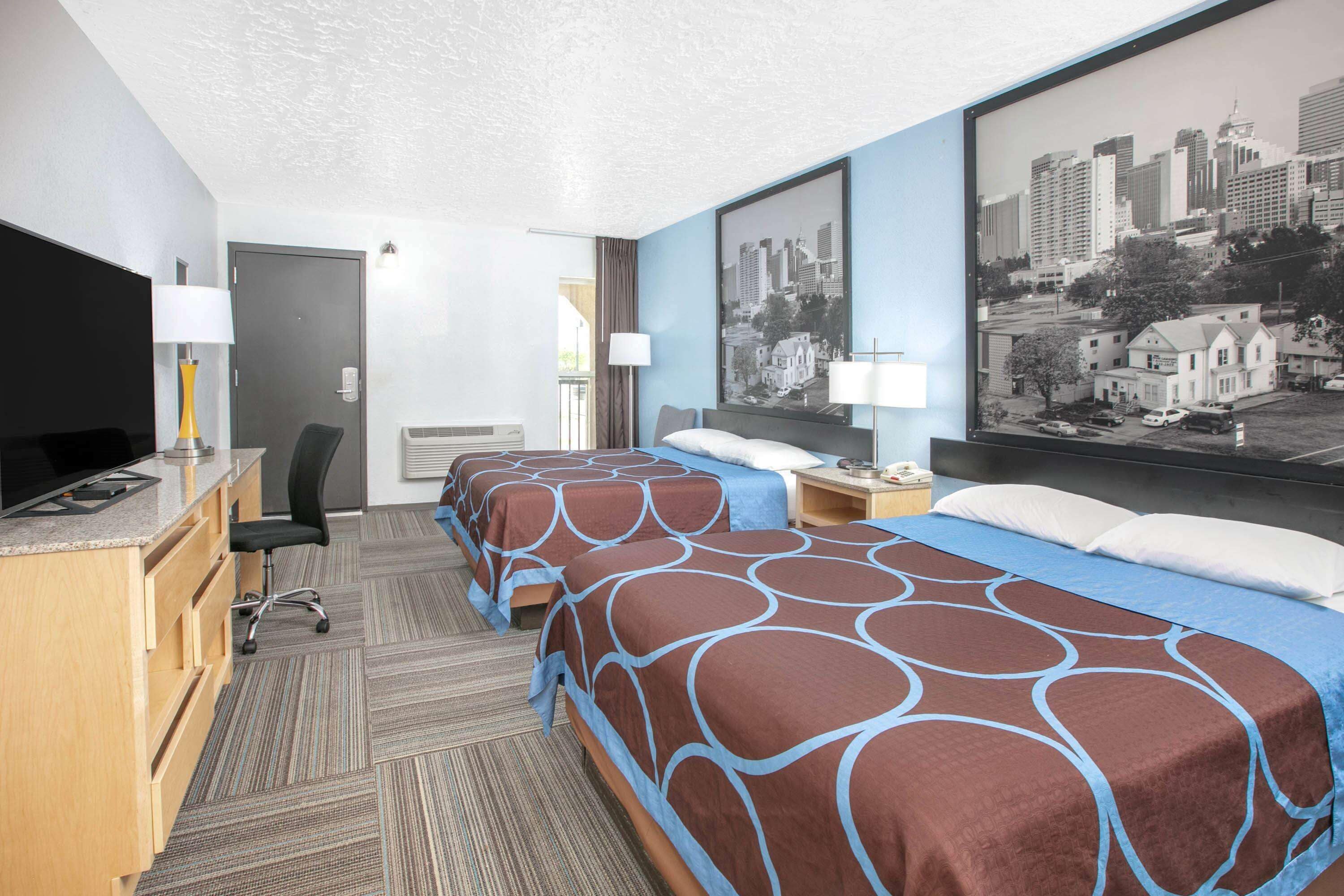A hotel room with two beds, a desk with a TV, and a cityscape painting on the wall.