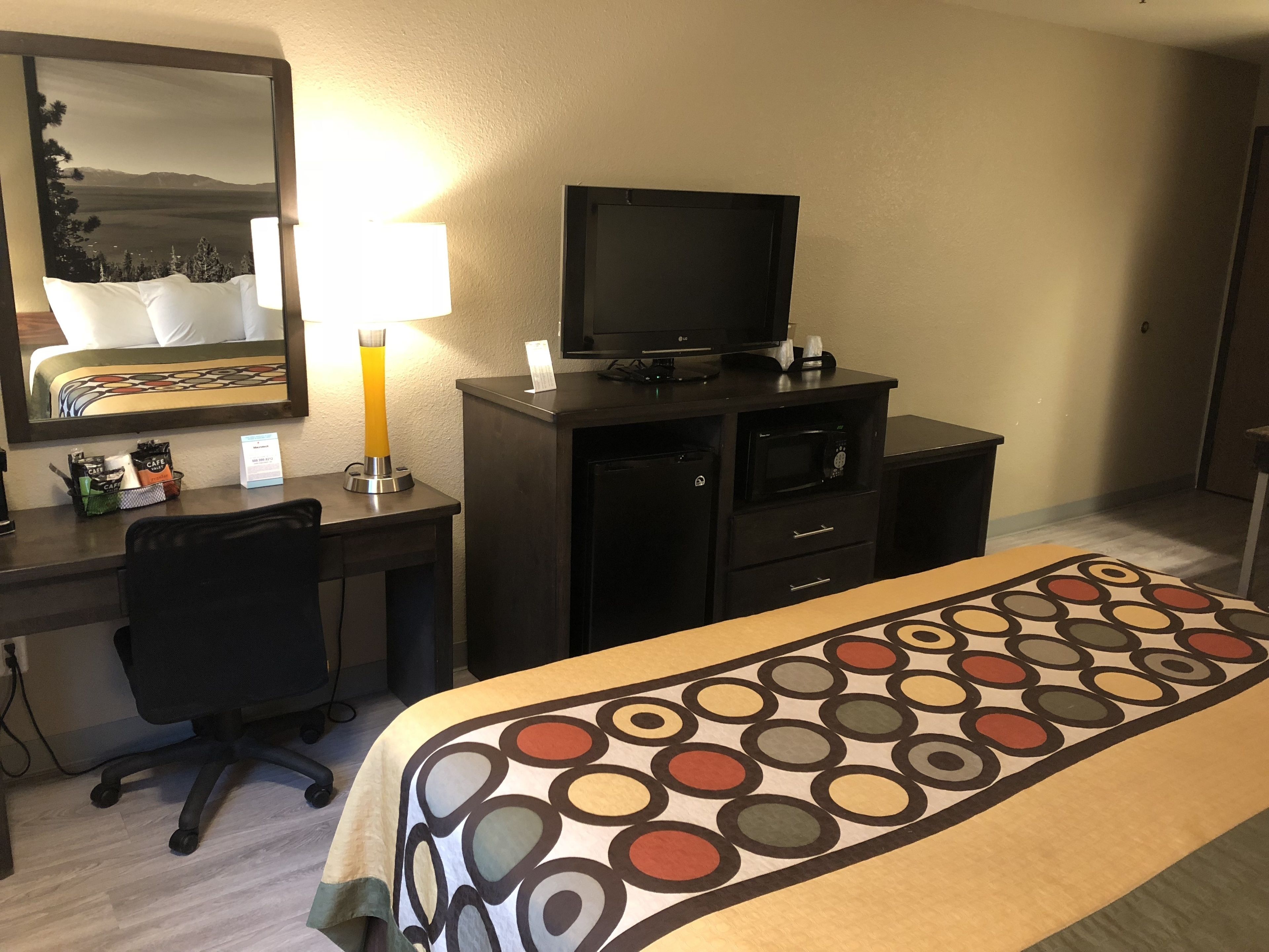 Habitación de hotel con cama, escritorio, silla, televisor, lámpara y cafetera.
