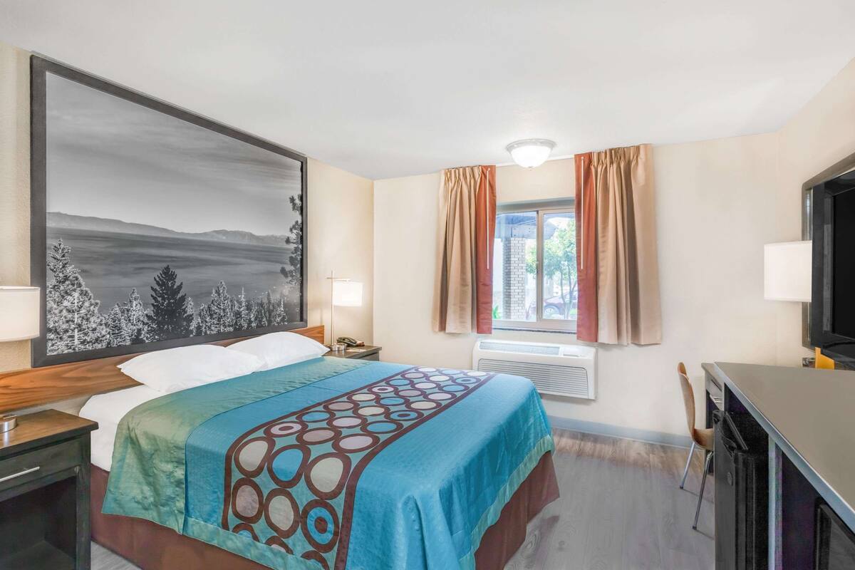 A hotel room with a bed, a desk, a television, a window with curtains, and a painting on the wall.