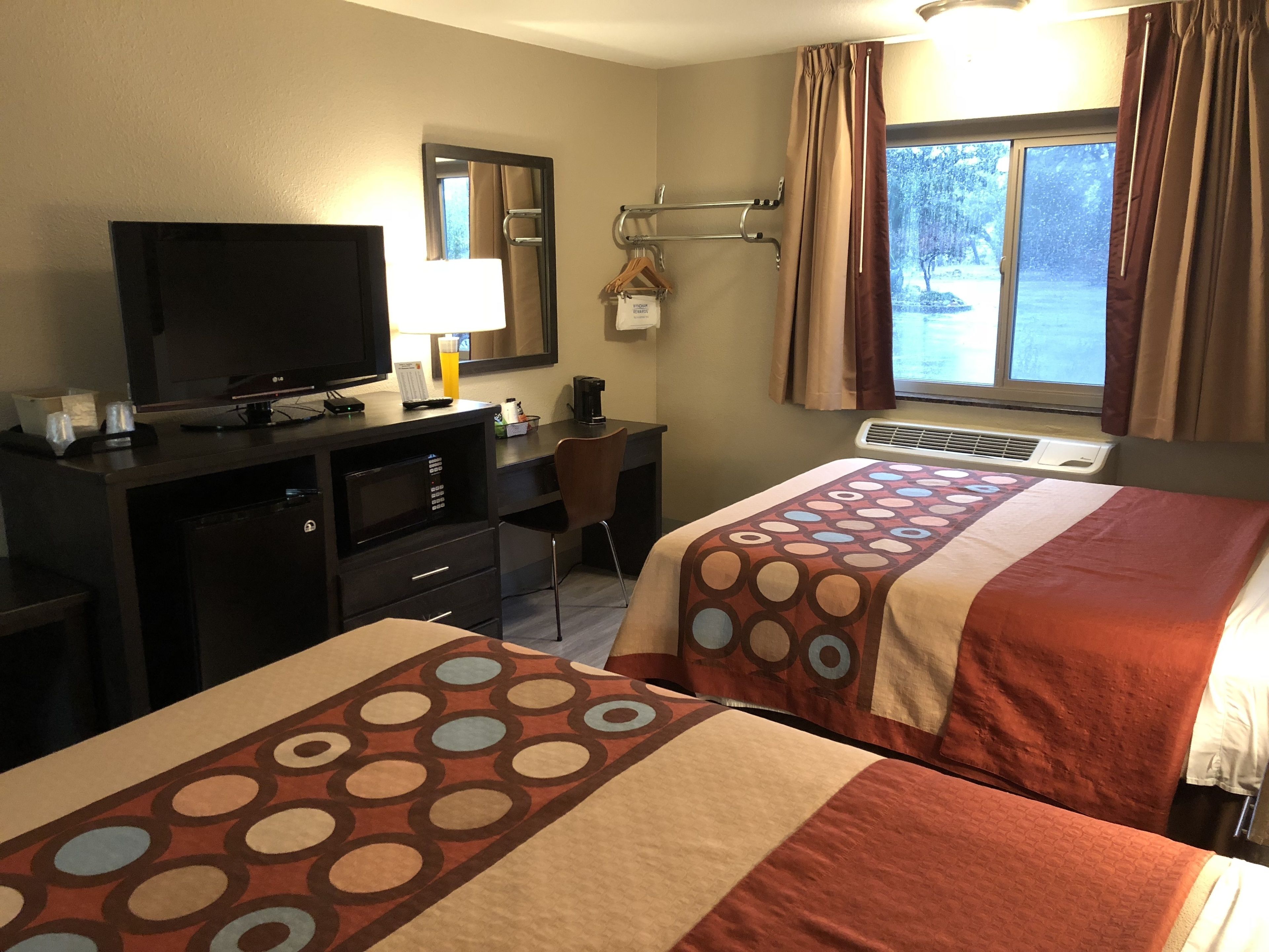 Quarto de hotel com duas camas, uma escrivaninha, televisão, luminária e janela com cortinas.