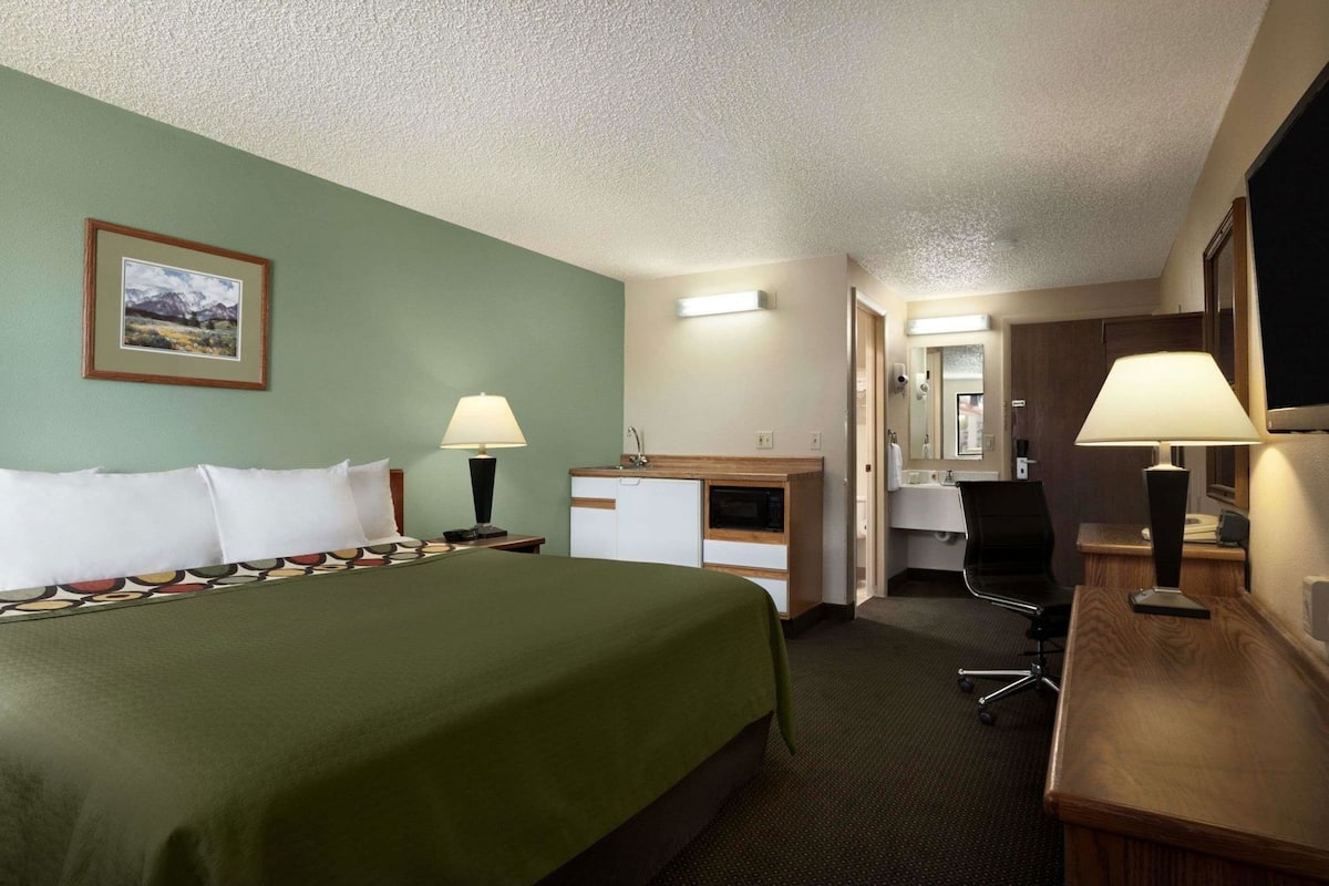 A hotel room with a bed, a desk, a chair, a desk lamp, a sink, and a mirror.