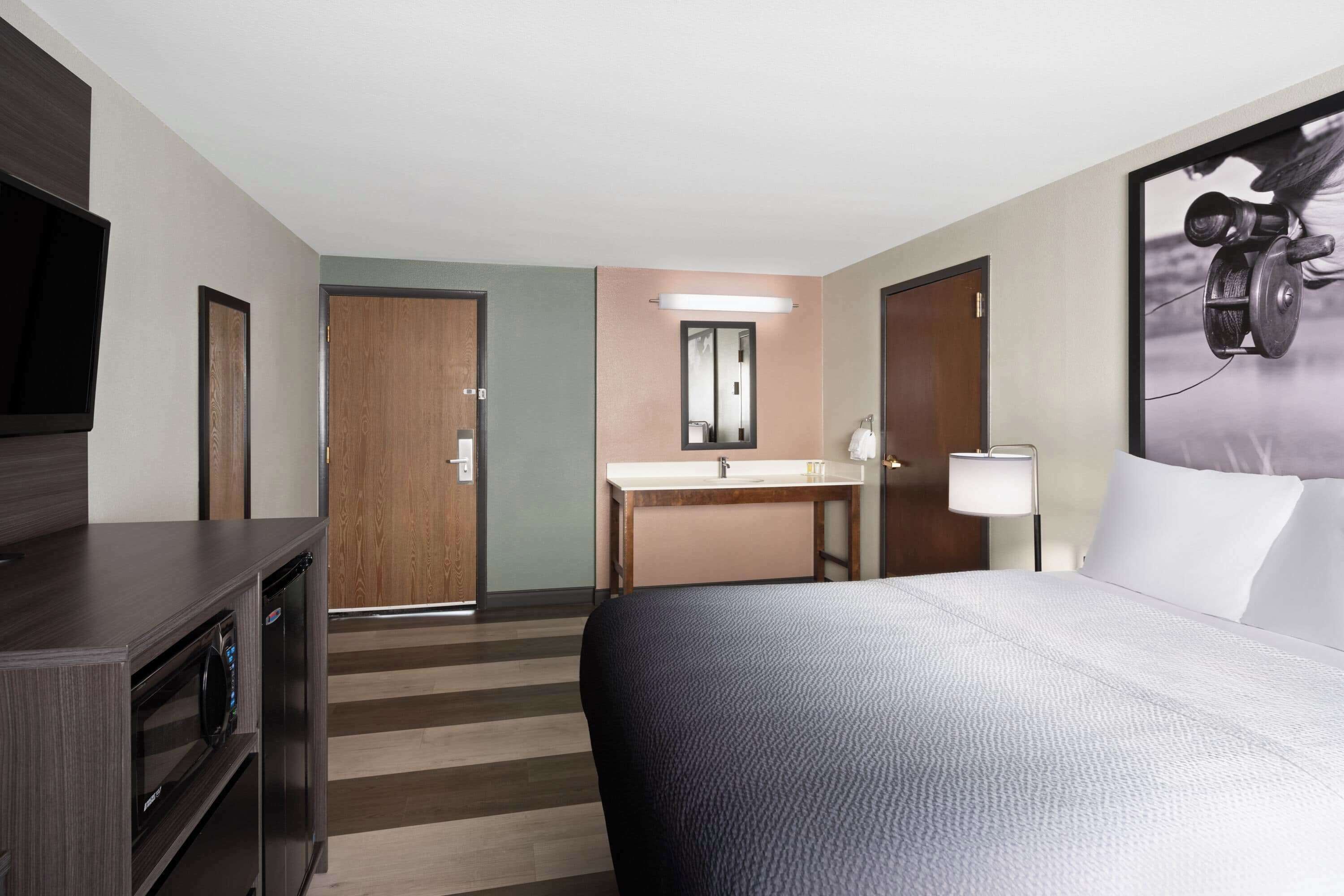 A hotel room with a bed, a desk, a TV, a bathroom with a sink and mirror, and a door.
