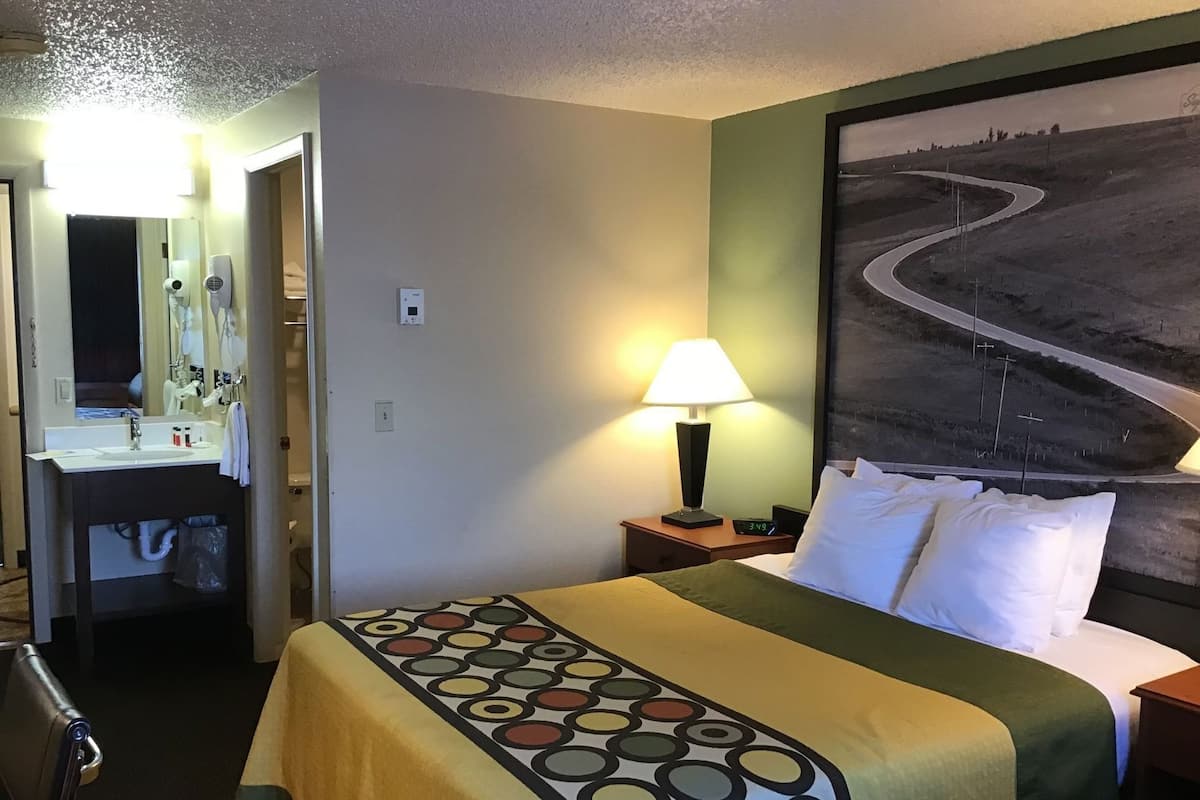 A hotel room with a bed, a nightstand with a lamp, a painting of a road on the wall, and a bathroom visible in the background.