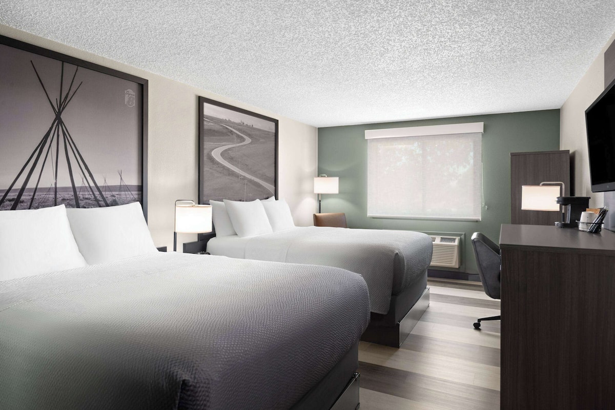 A hotel room with two beds, a desk, a lamp, and a large window.