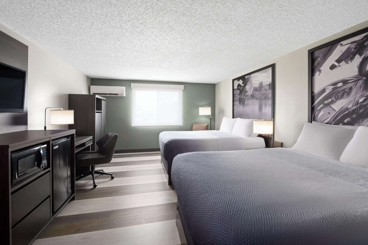 A hotel room with two beds, a desk with a microwave, a chair, a TV, and artwork on the walls.