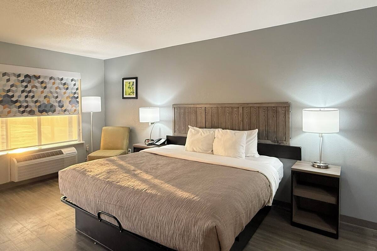 Una habitación de hotel con una cama grande, dos mesitas de noche con lámparas, una silla, un calentador de pared y un adorno de pared con estampado geométrico.