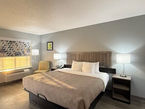 A hotel room with a large bed, two bedside tables with lamps, a chair, a wall-mounted heater, and a geometric-patterned wall decoration. - Quality Inn Crestview I-10 Eglin AFB Area (Crestview)