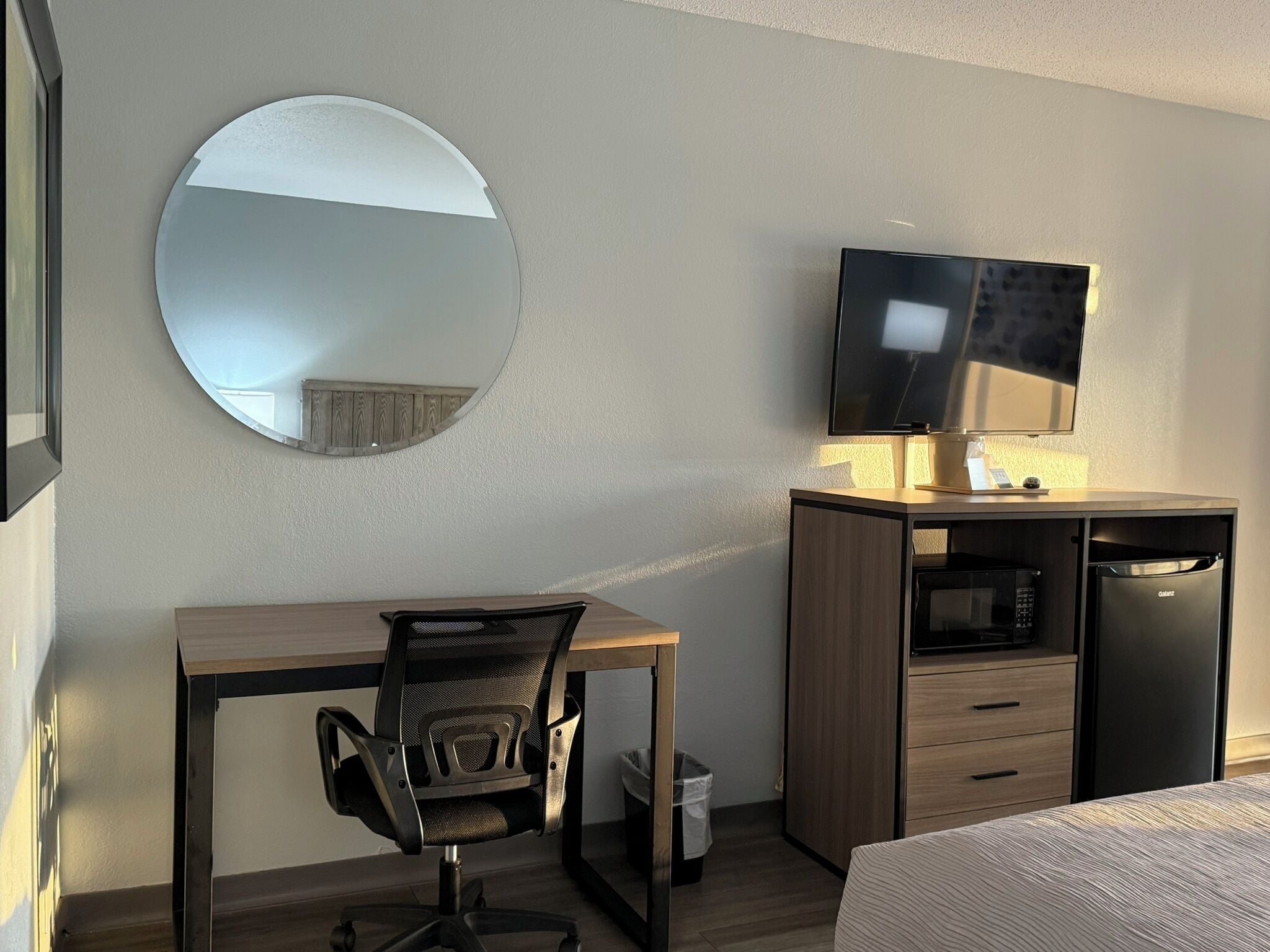 Ett hotellrum med ett skrivbord i trä, en svart kontorsstol, en rund spegel, en tv, en minibar och en säng.