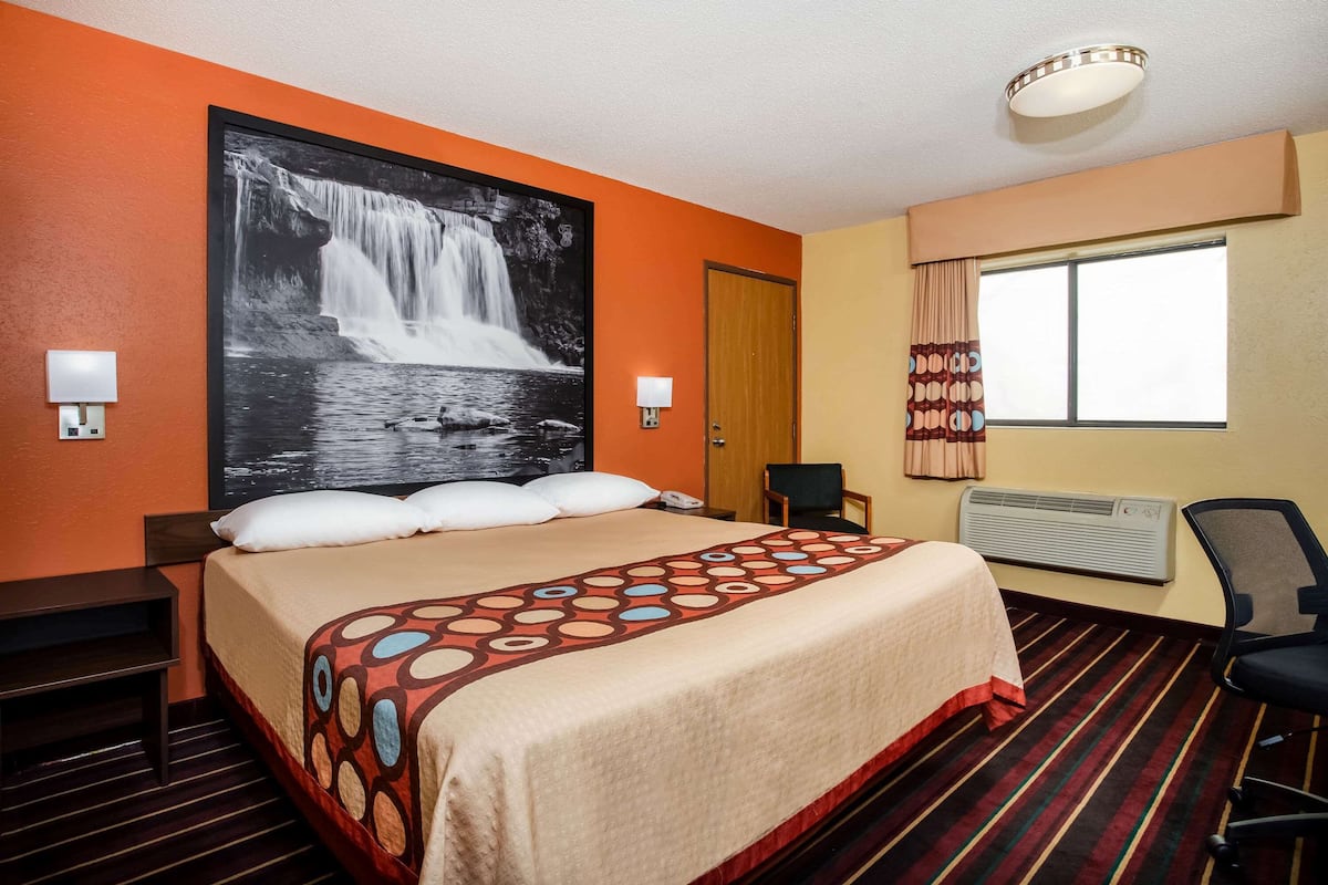 Ein Hotelzimmer mit einem großen Bett, einem an der Wand montierten Fernseher, einem Fenster mit Vorhängen und einem Schreibtisch mit Stuhl.